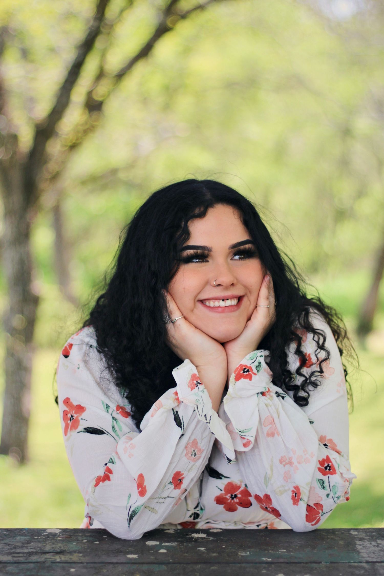 Cheerful outdoor portrait in floral white blouse resting chin on hands.