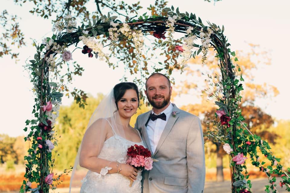 Newlyweds under floral arch during outdoor sunset wedding ceremony.