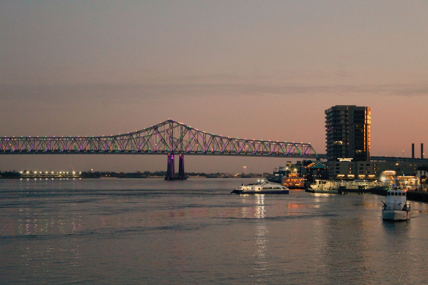 Glowing bridge lights reflect on calm water during sunset creating a peaceful panoramic cityscape scene.