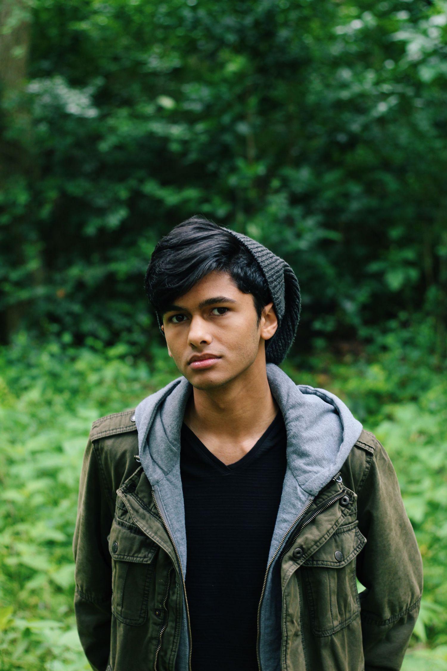 A moody portrait in an olive jacket and beanie against lush green forest background.