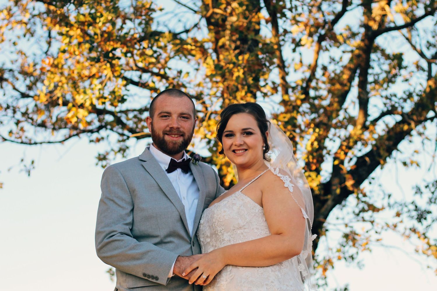 A couple poses outdoors on their wedding day with autumn foliage in the background.