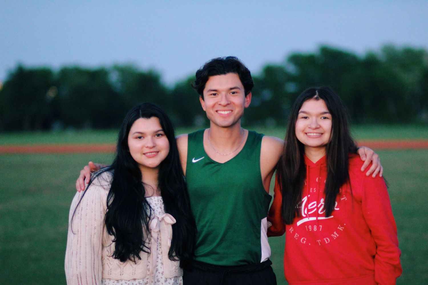 Three siblings posing together outdoors on a grassy field with trees in background.