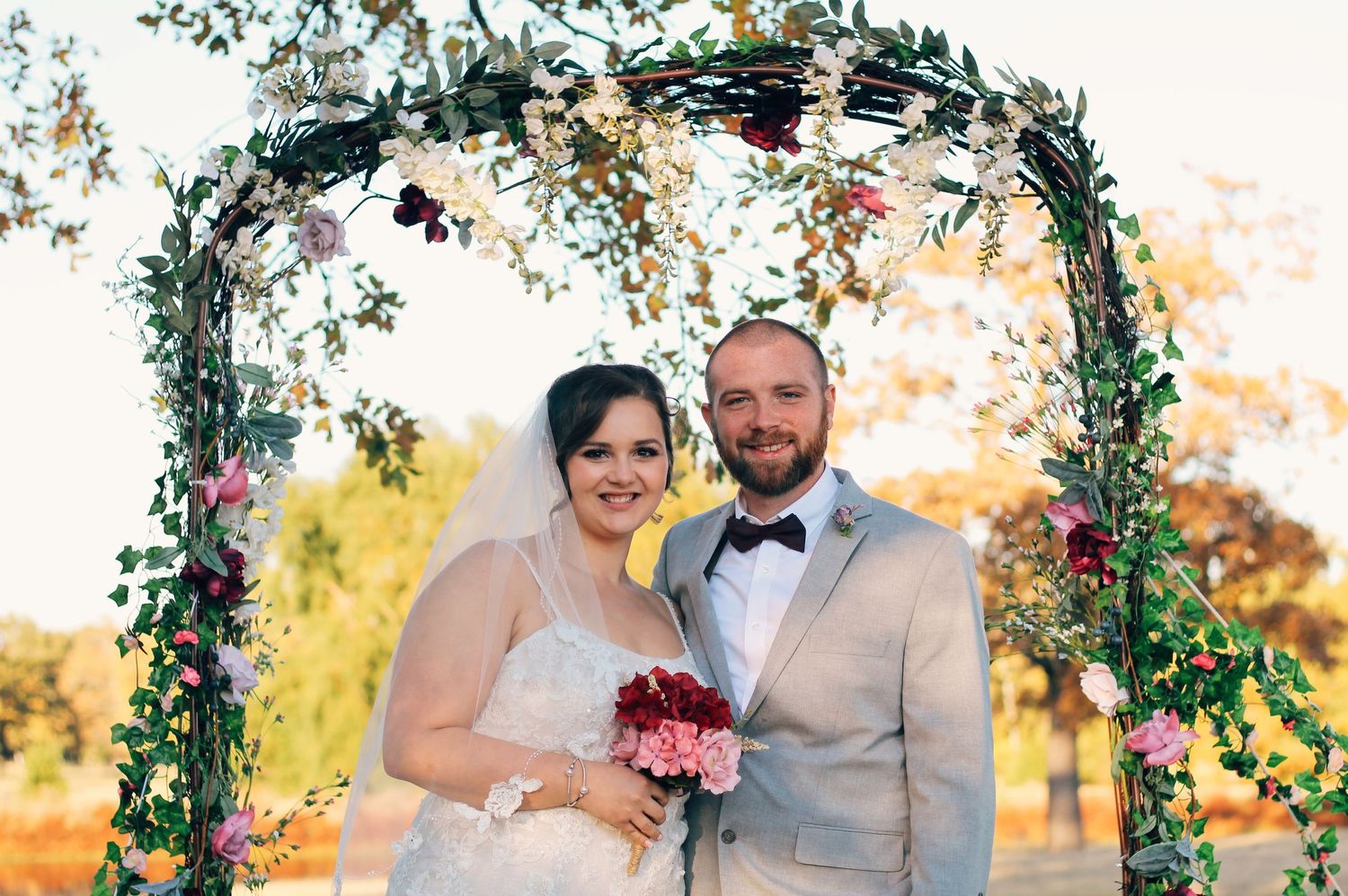 Happy couple poses under decorated floral arch at their autumn wedding.