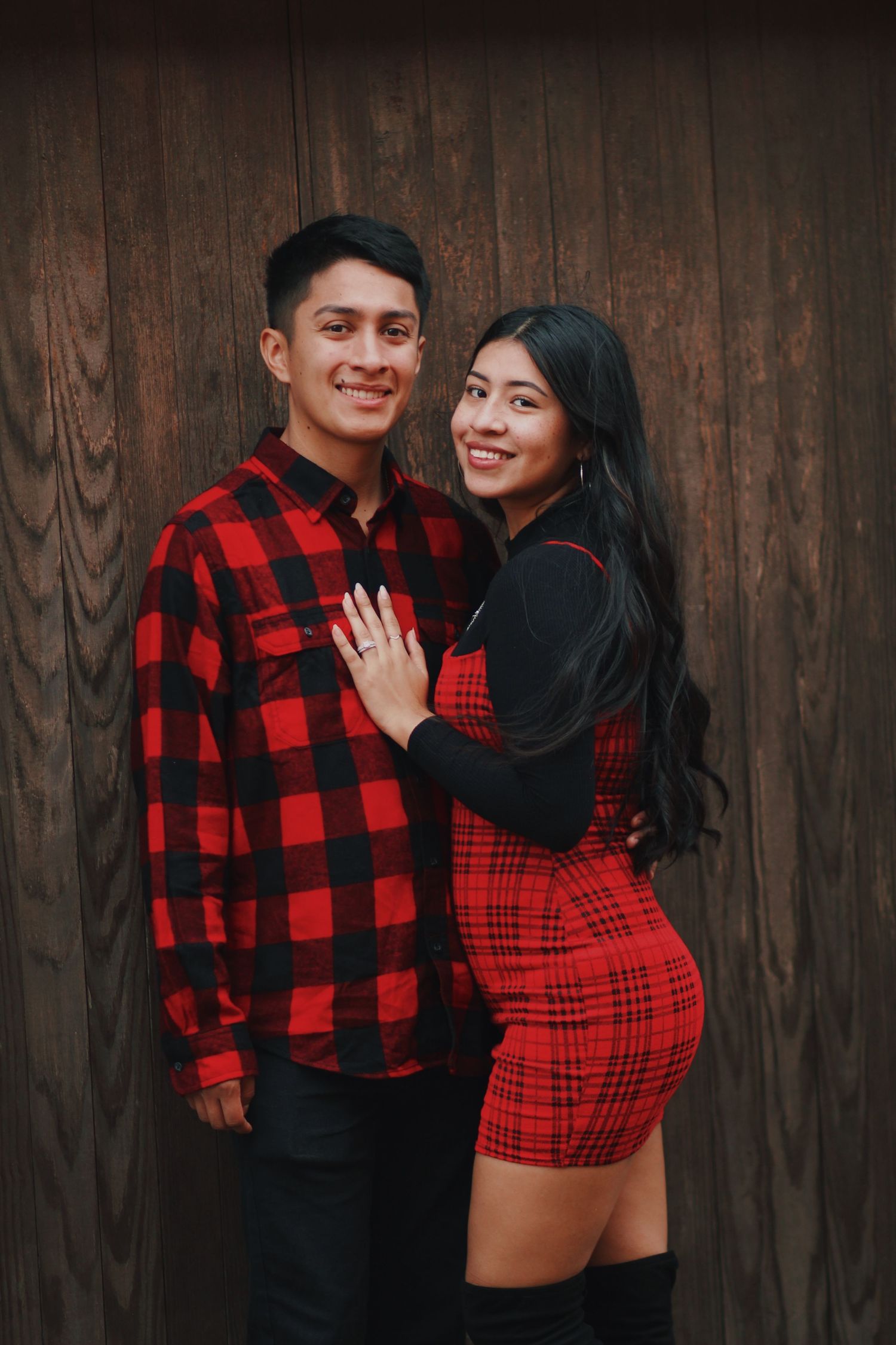 A couple in coordinating red and black outfits poses against a wooden wall backdrop.