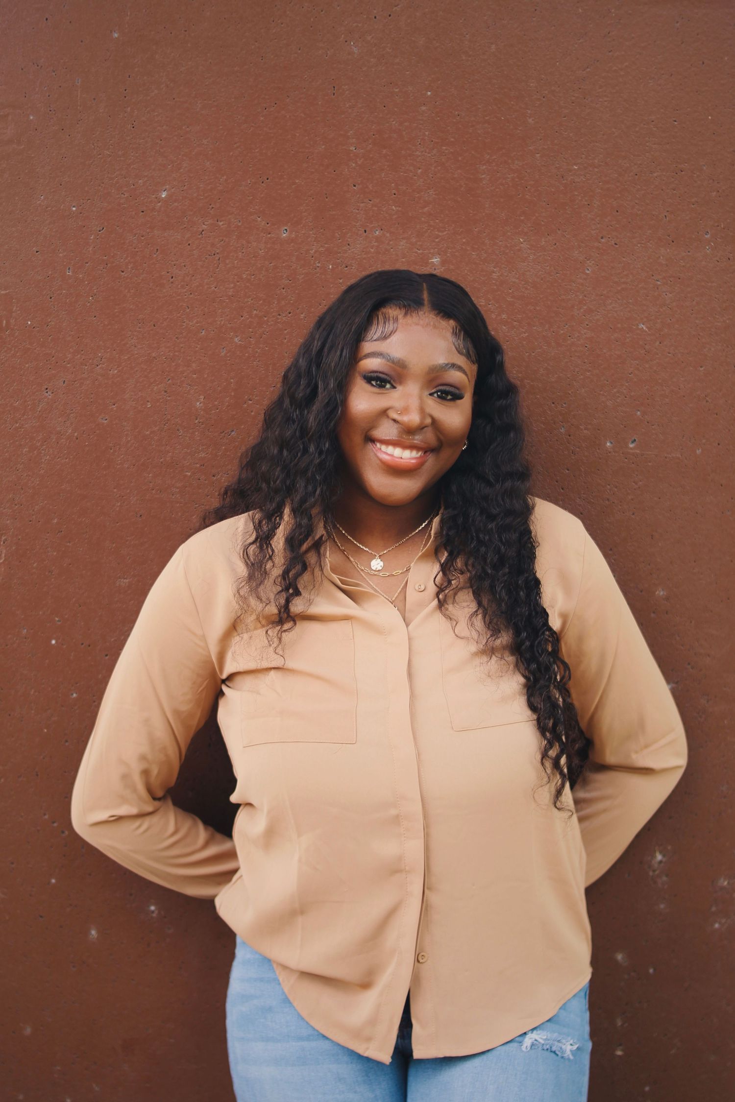 Portrait against brown wall shows warm smile and long curly hair with beige button-up and distressed jeans.