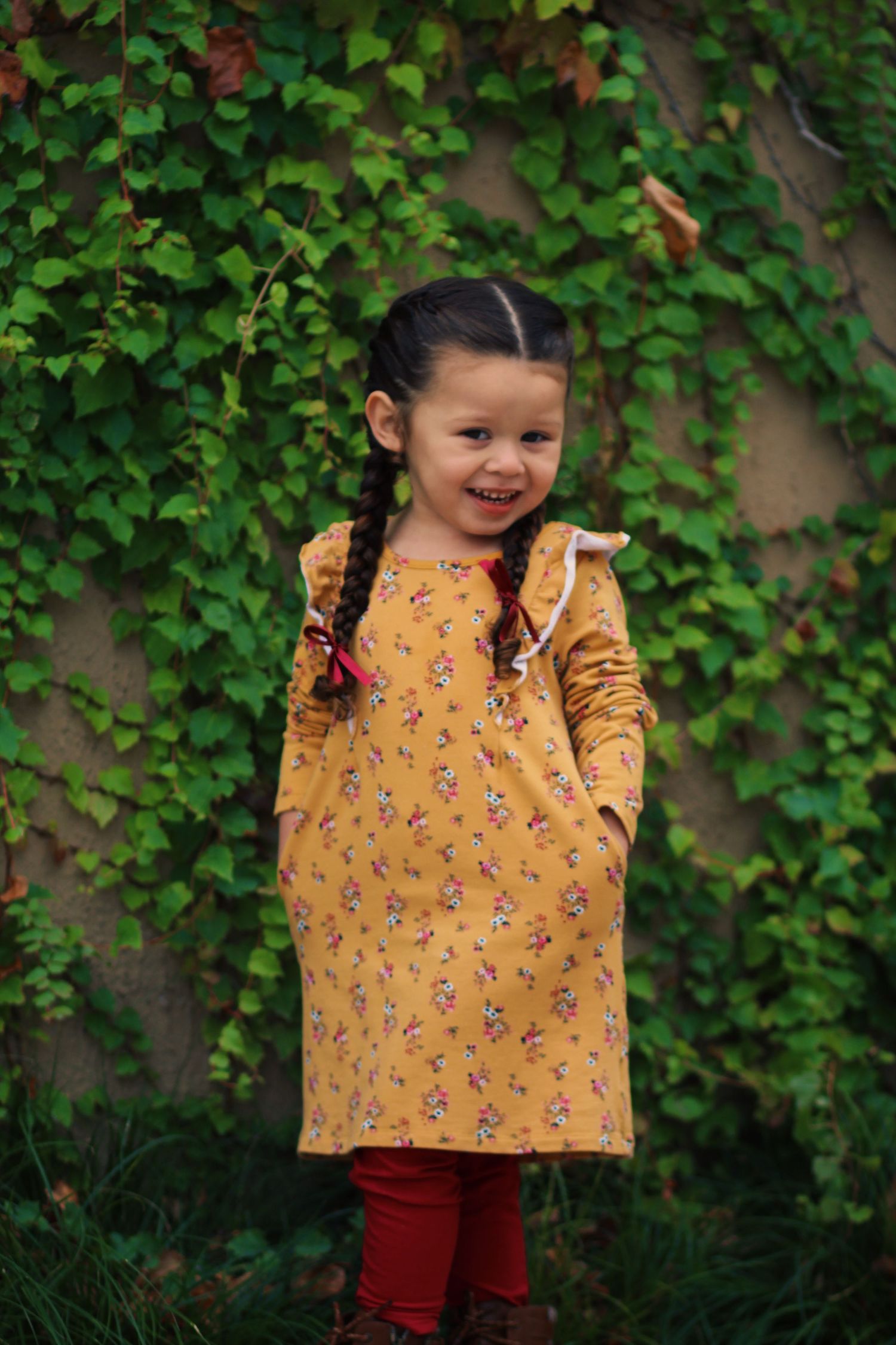 An energetic pose in a yellow floral dress against lush green ivy backdrop.