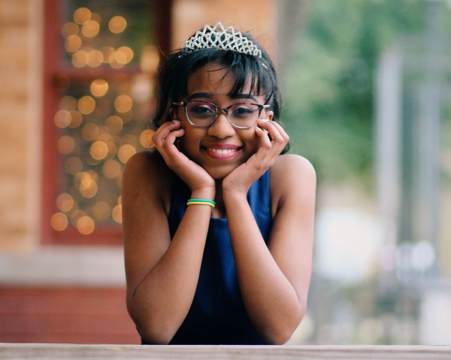 A person wearing a tiara and navy dress poses cheerfully with hands framing their face.