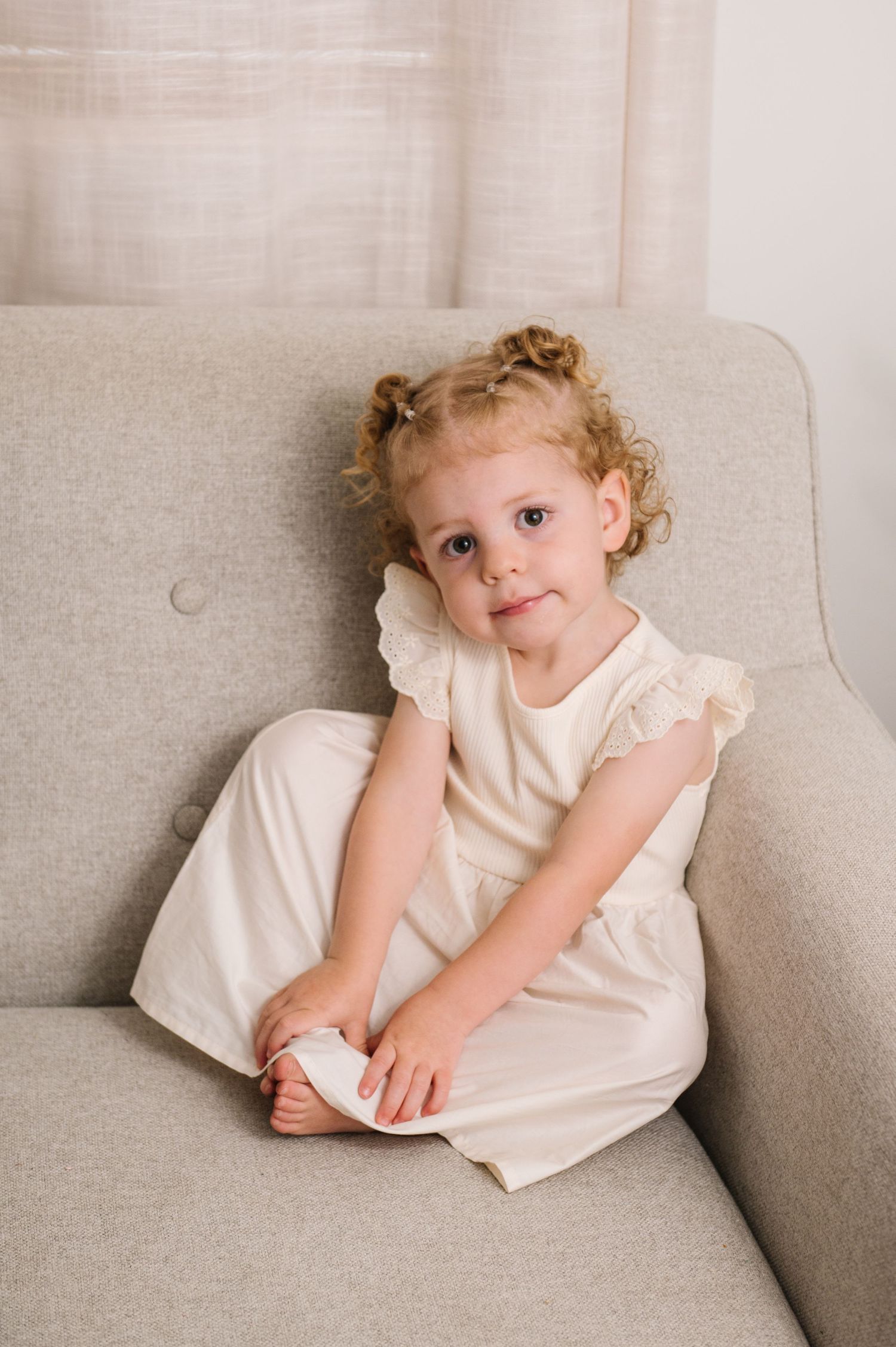 Young child in white dress sitting on grey couch with curly hair.