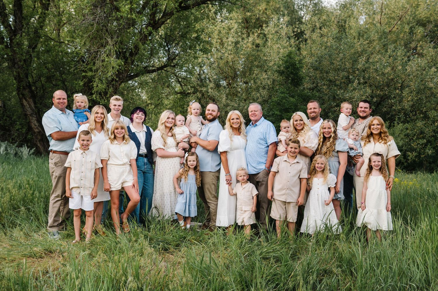 Large group portrait outdoors in coordinated blue and beige outfits standing together in tall grass with trees behind them.