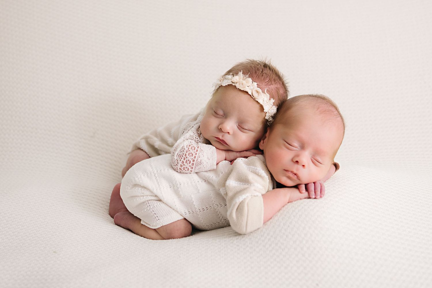 Two small babies in white outfits sleeping peacefully on a light backdrop.