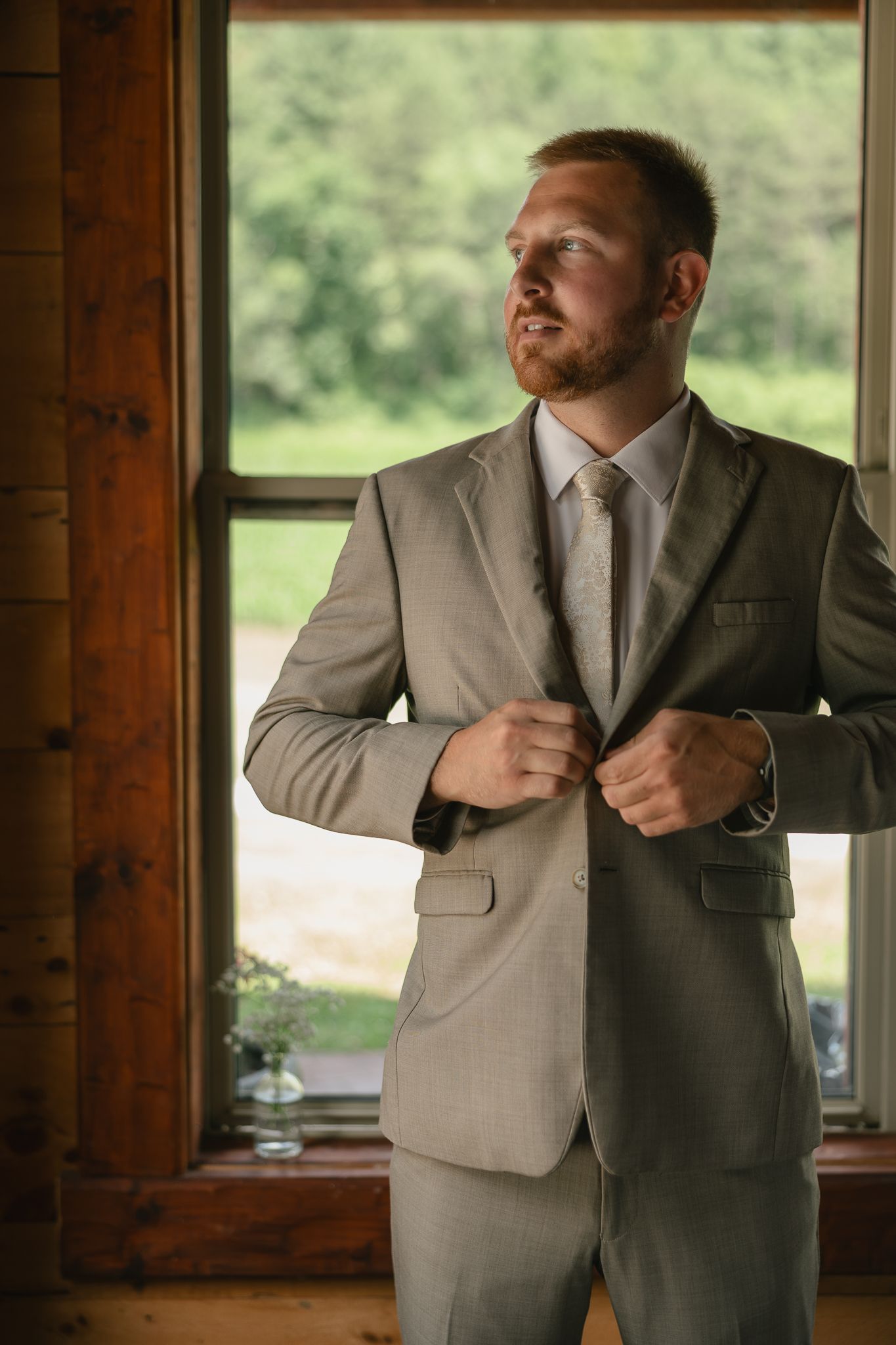 Man in light gray suit preparing for wedding in wooden cabin.