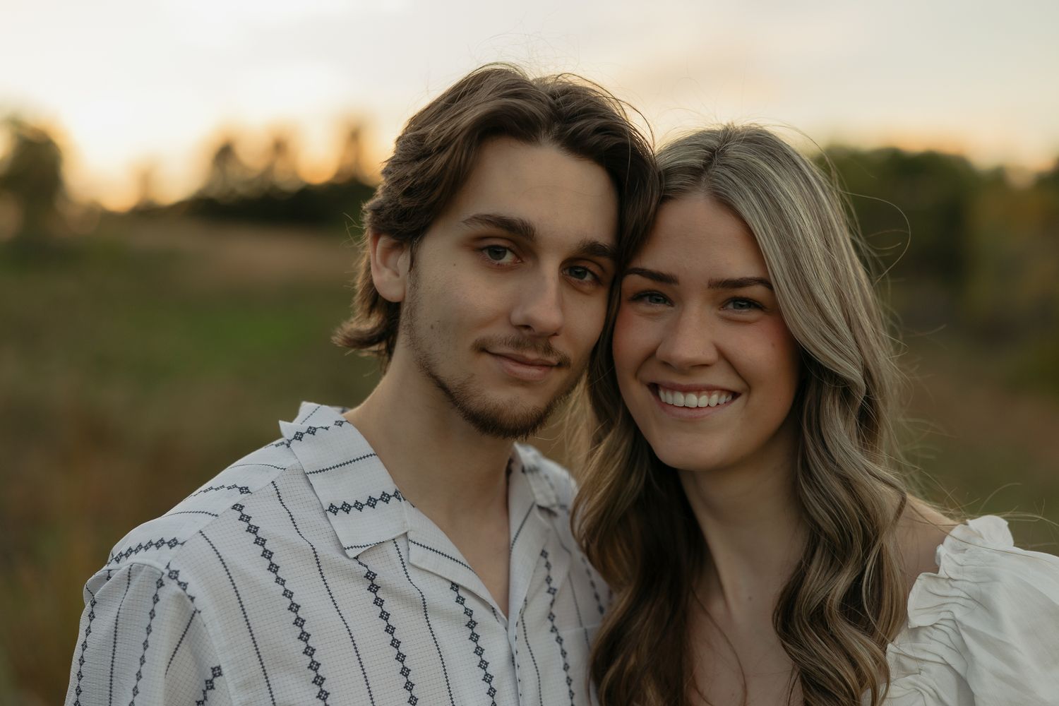 A couple smiles warmly at each other in a golden hour outdoor setting.