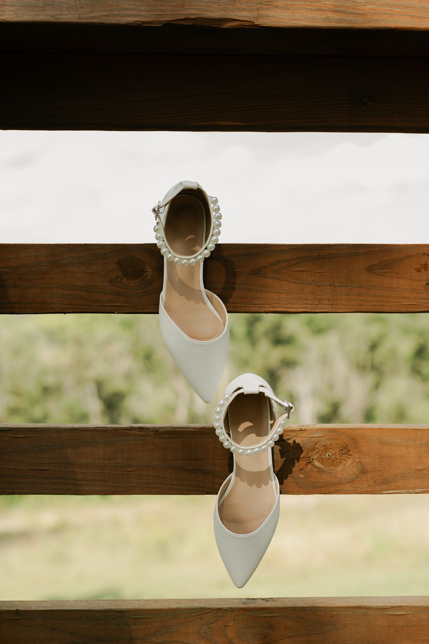 Elegant white bridal shoes with pearl details displayed on wooden beams.