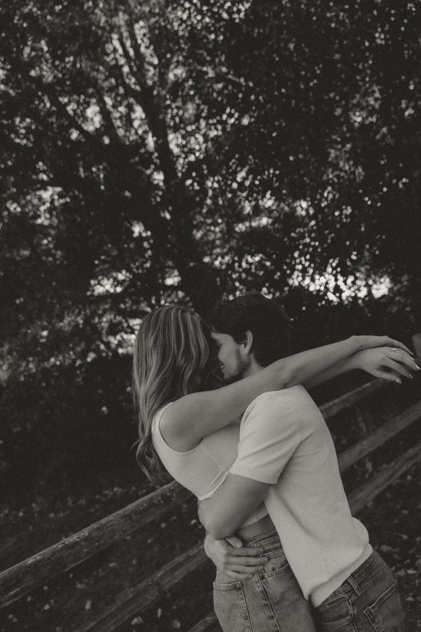 Couple embracing tenderly against rustic wooden fence backdrop in black and white photography.
