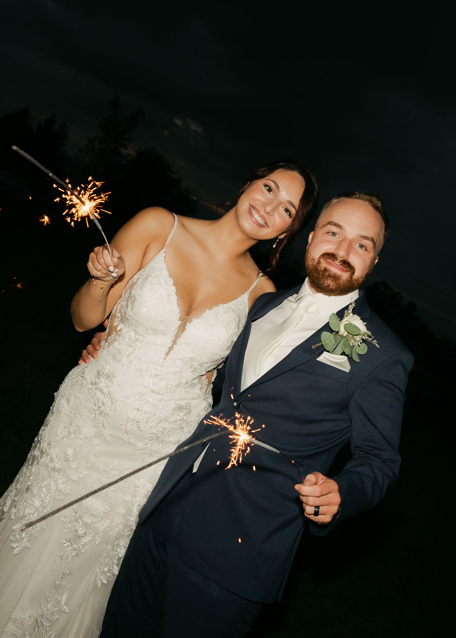 Newlyweds smile while holding sparklers during evening wedding festivities.