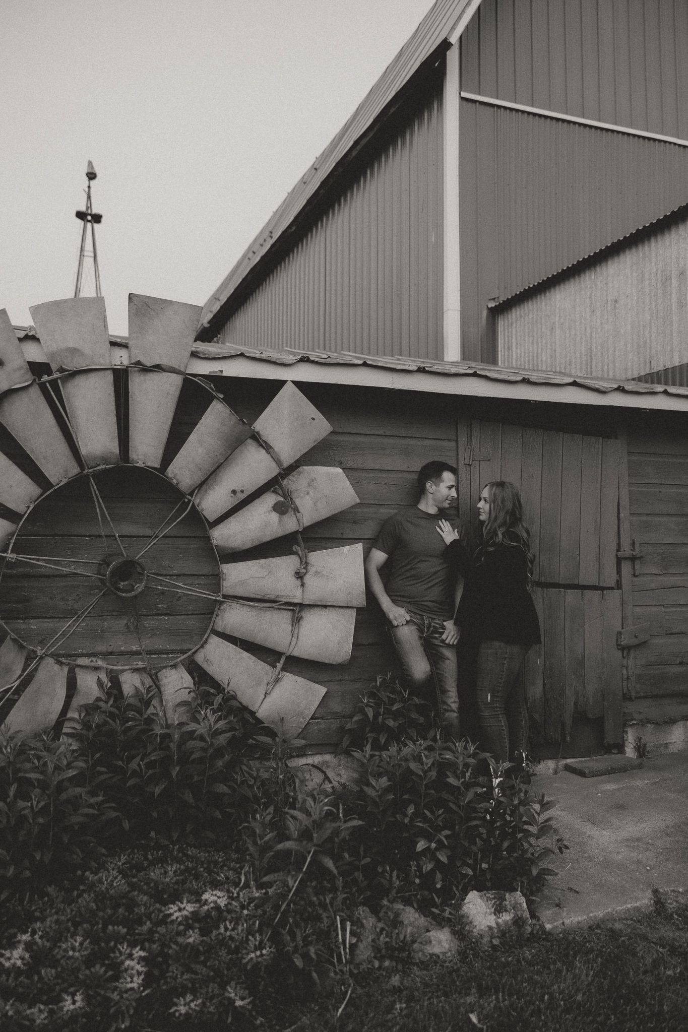 A couple poses beside a vintage windmill wheel against a rustic barn wall.