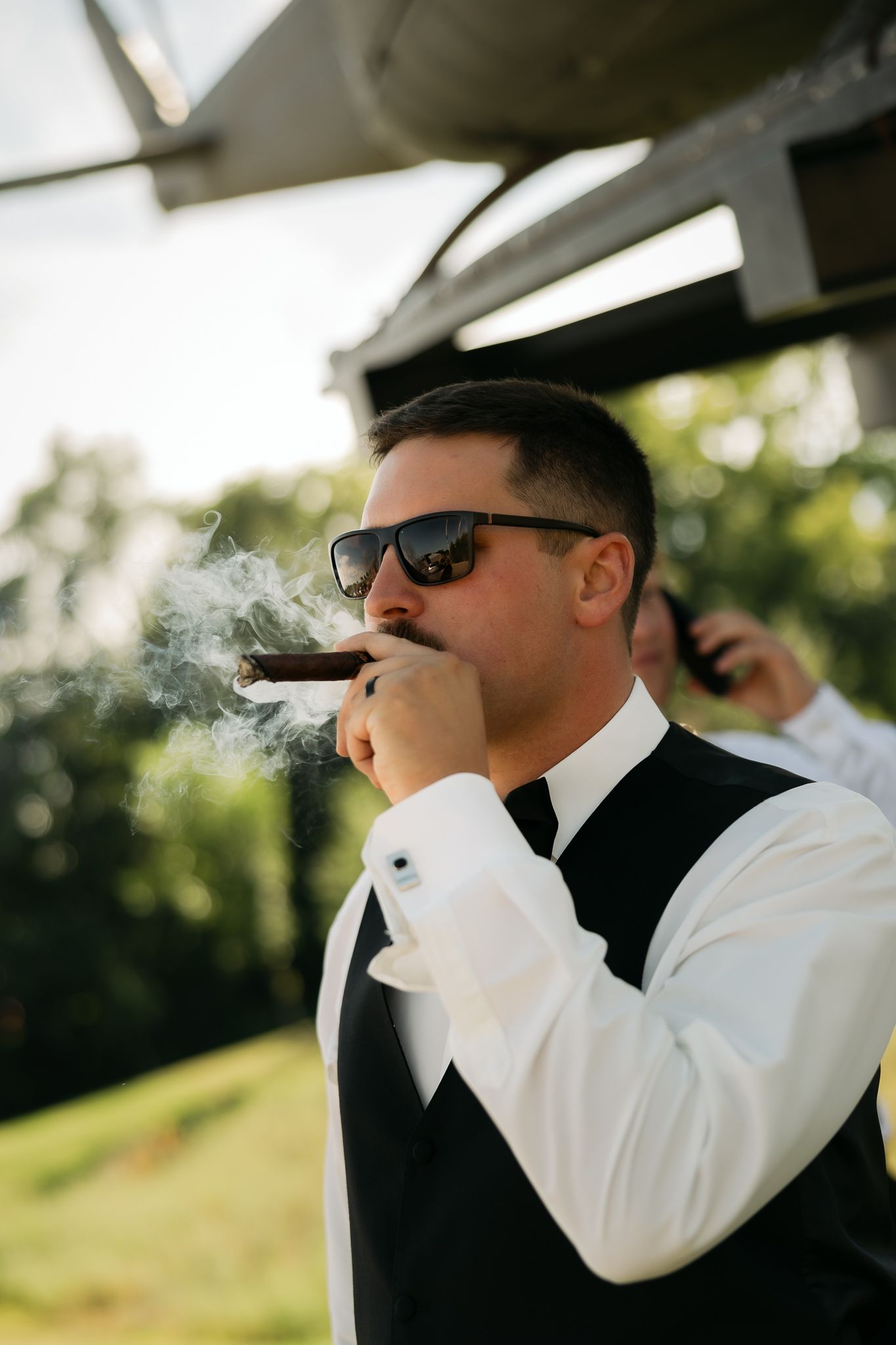 A person in formal attire and sunglasses smoking a cigar outdoors on a sunny day.