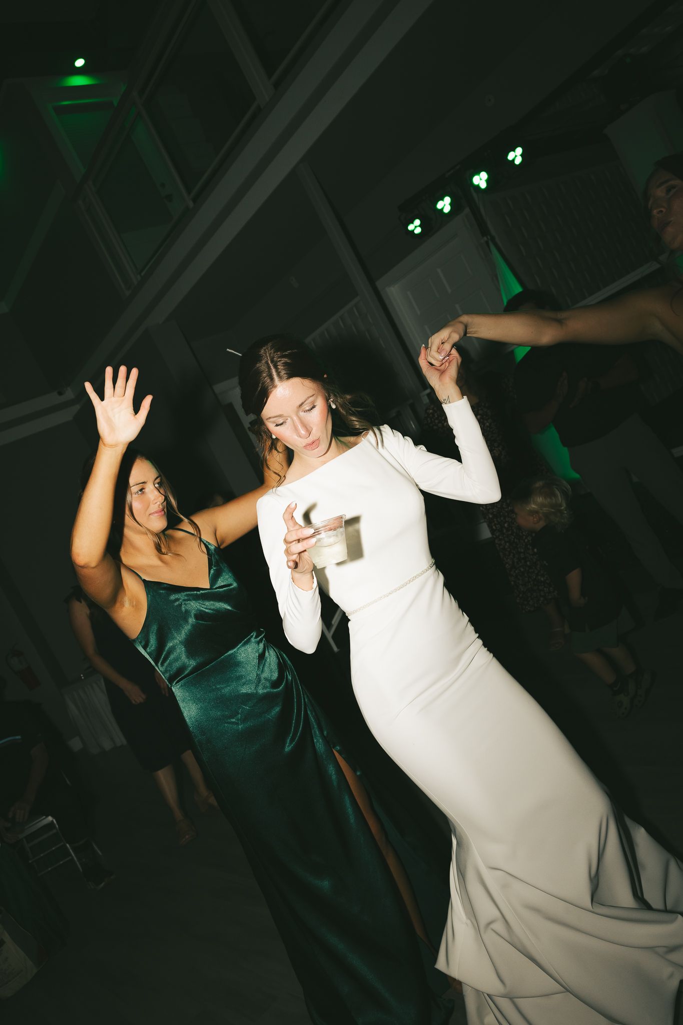 Two women in elegant dresses dancing energetically at a reception with green lighting.