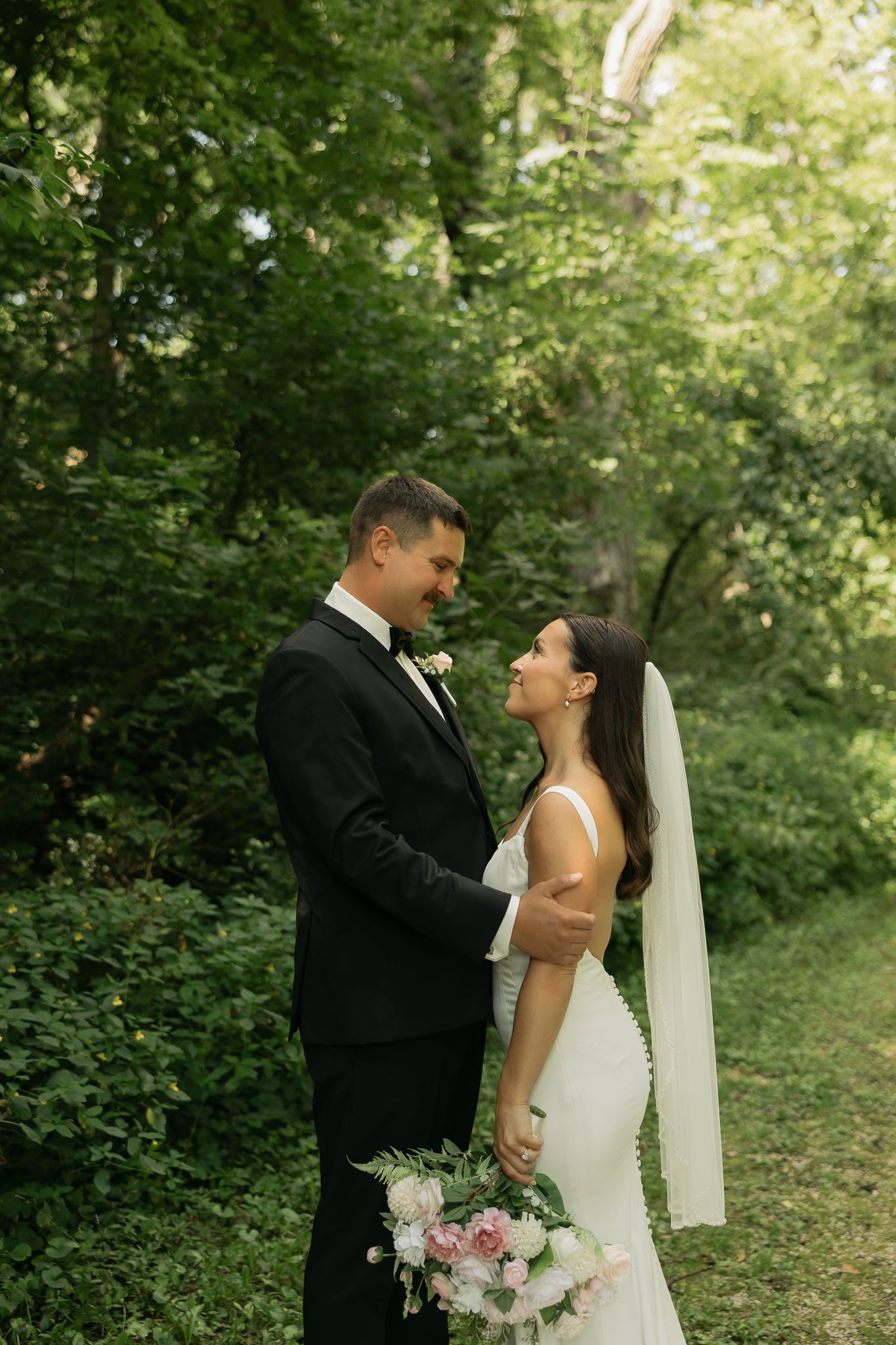 Intimate wedding portrait of a couple in formal attire sharing a tender moment in a wooded setting.