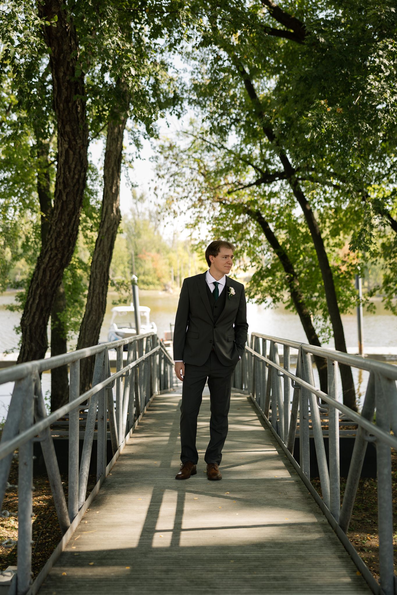 Groom in classic black suit stands on wooden bridge surrounded by lush trees.