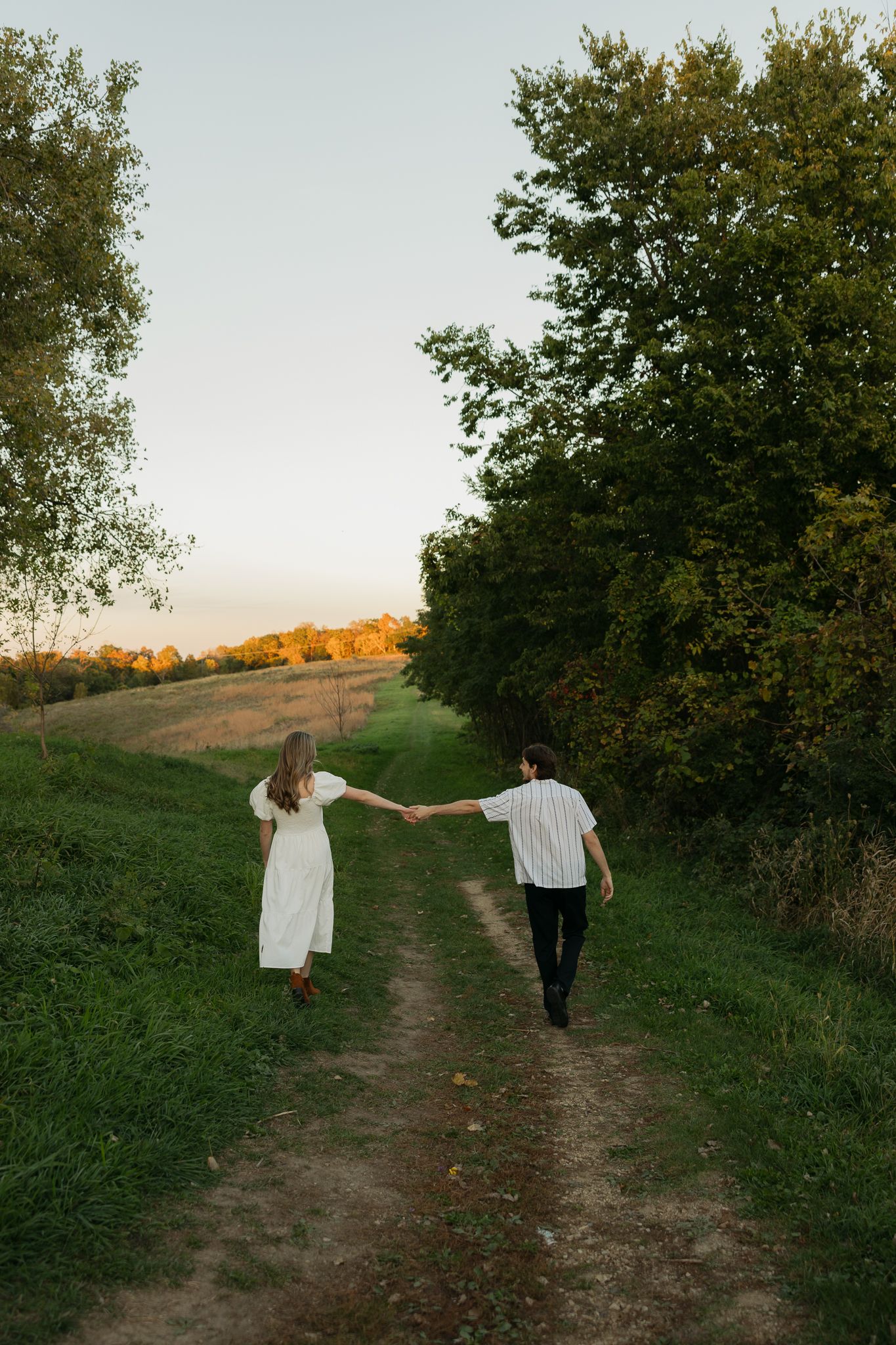 Couple walking hand in hand on dirt path during golden hour surrounded by trees.