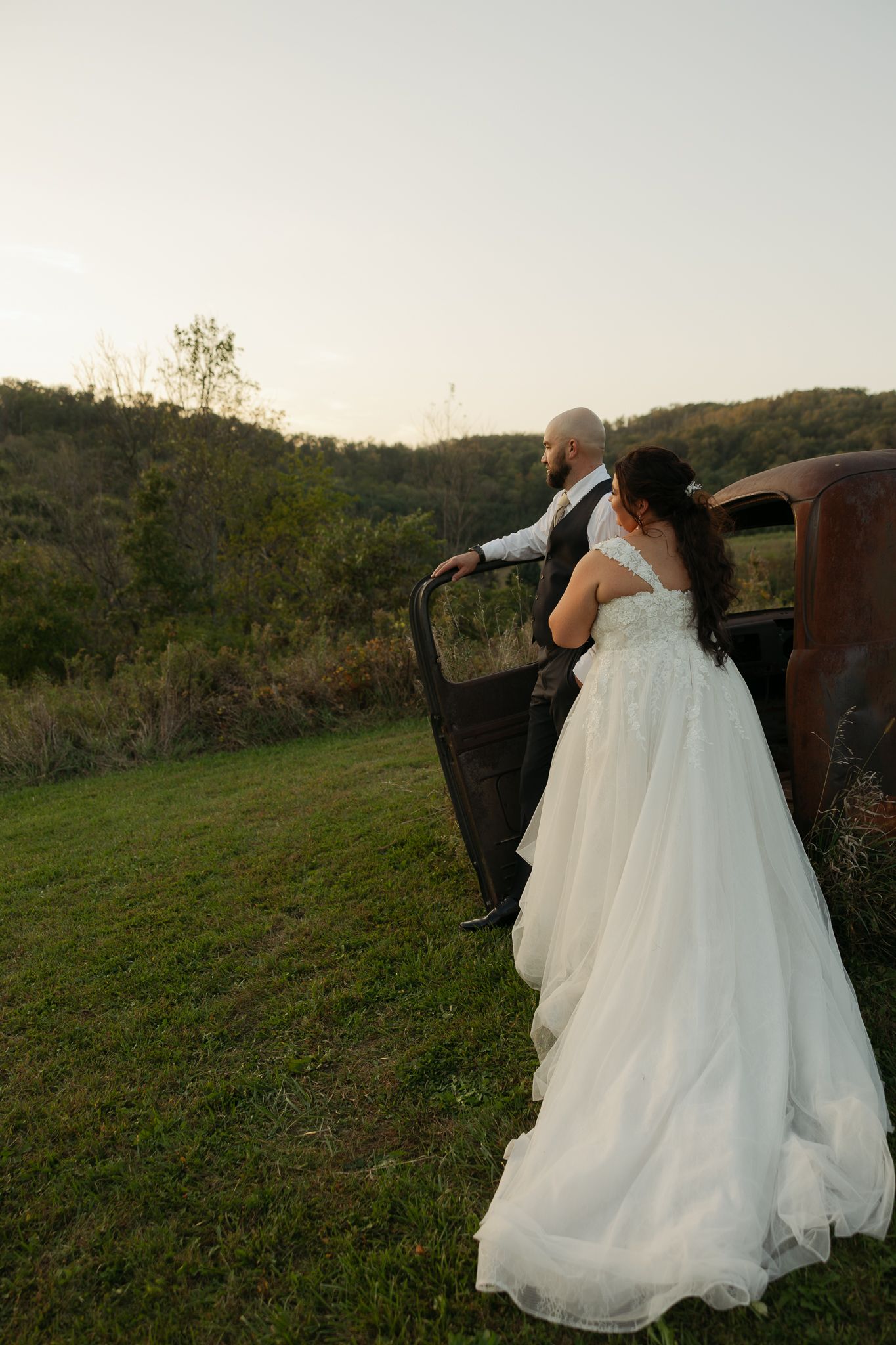 Wedding couple poses dramatically against sunset in flowing white gown.
