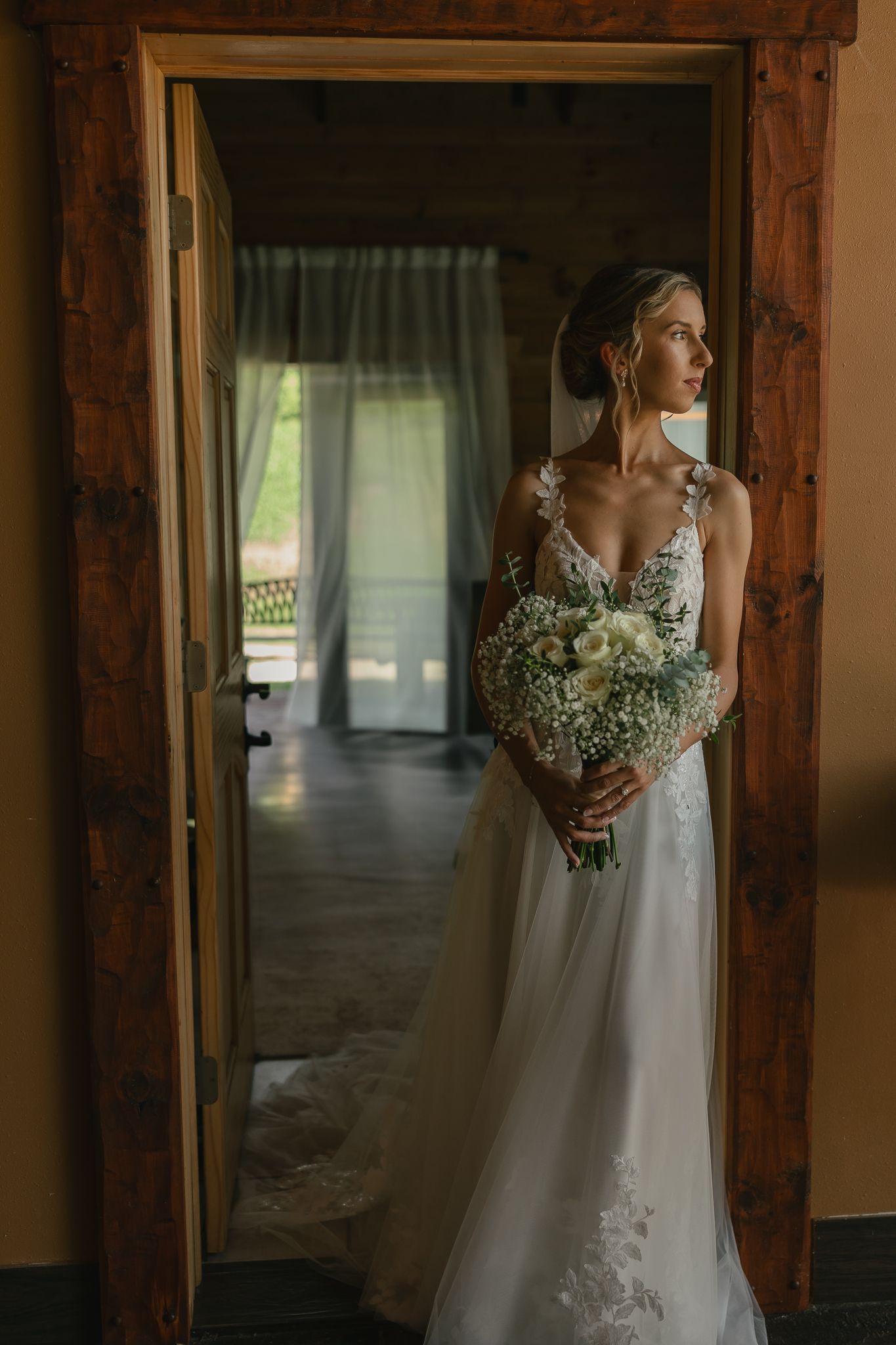 Bride in a white lace gown holding a bouquet poses near rustic wooden beams in natural light.