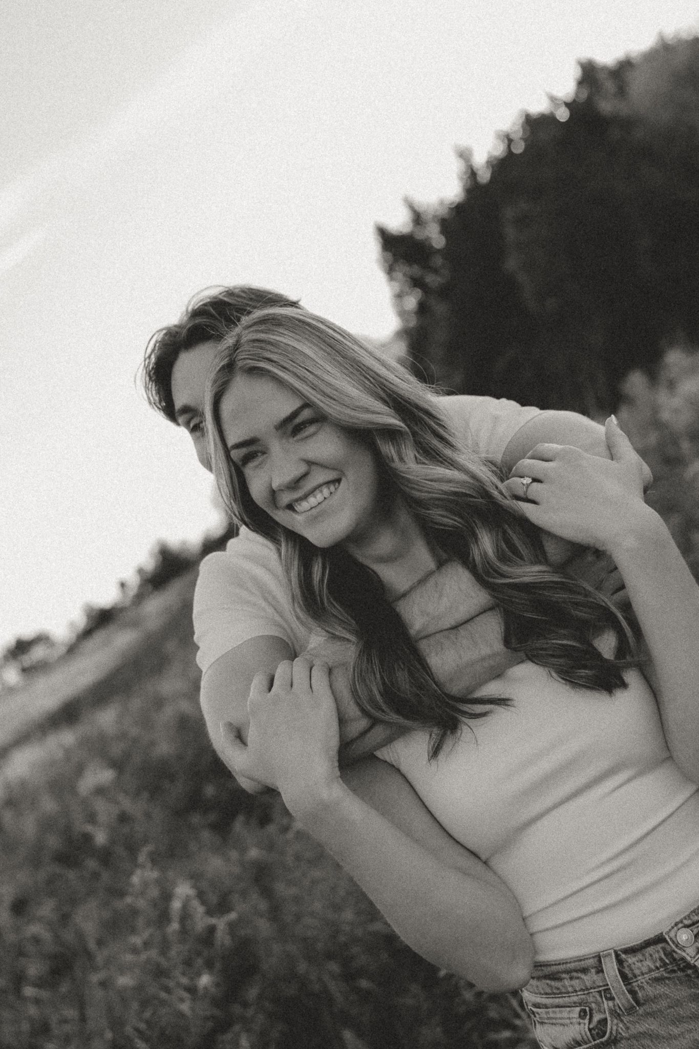 Black and white portrait of a smiling woman being embraced from behind in a natural setting.