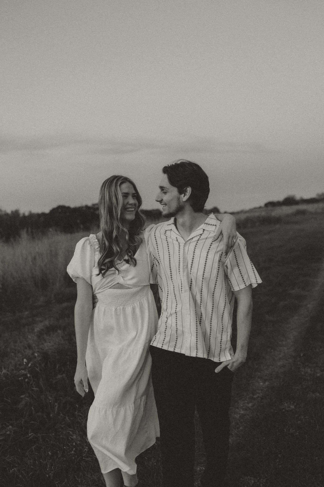 A couple in casual white outfits sharing intimate moments in a field at dusk.