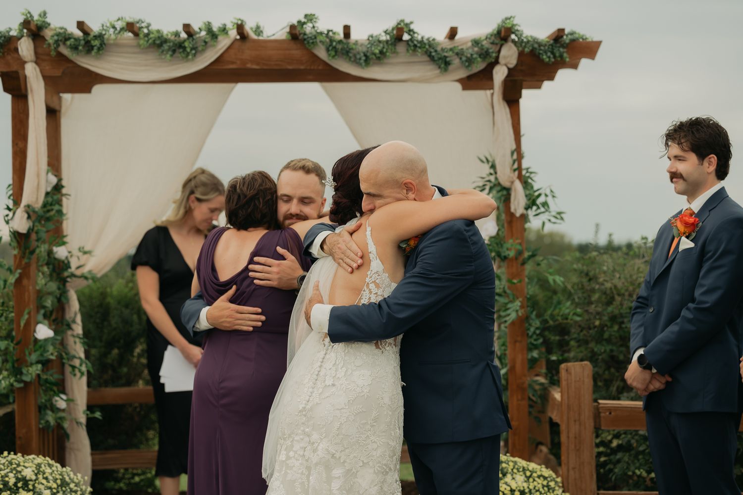 Outdoor wedding ceremony taking place under wooden arch adorned with greenery and fabric.