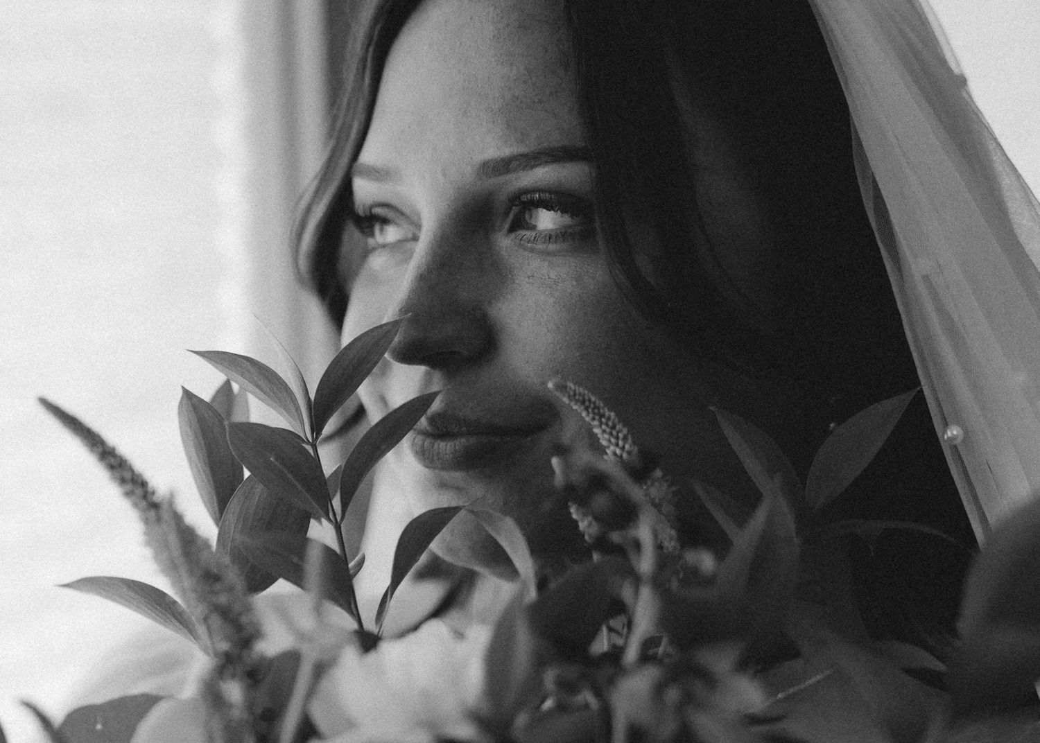 Black and white portrait of a bride holding a bouquet with a veil, gazing thoughtfully near a window.