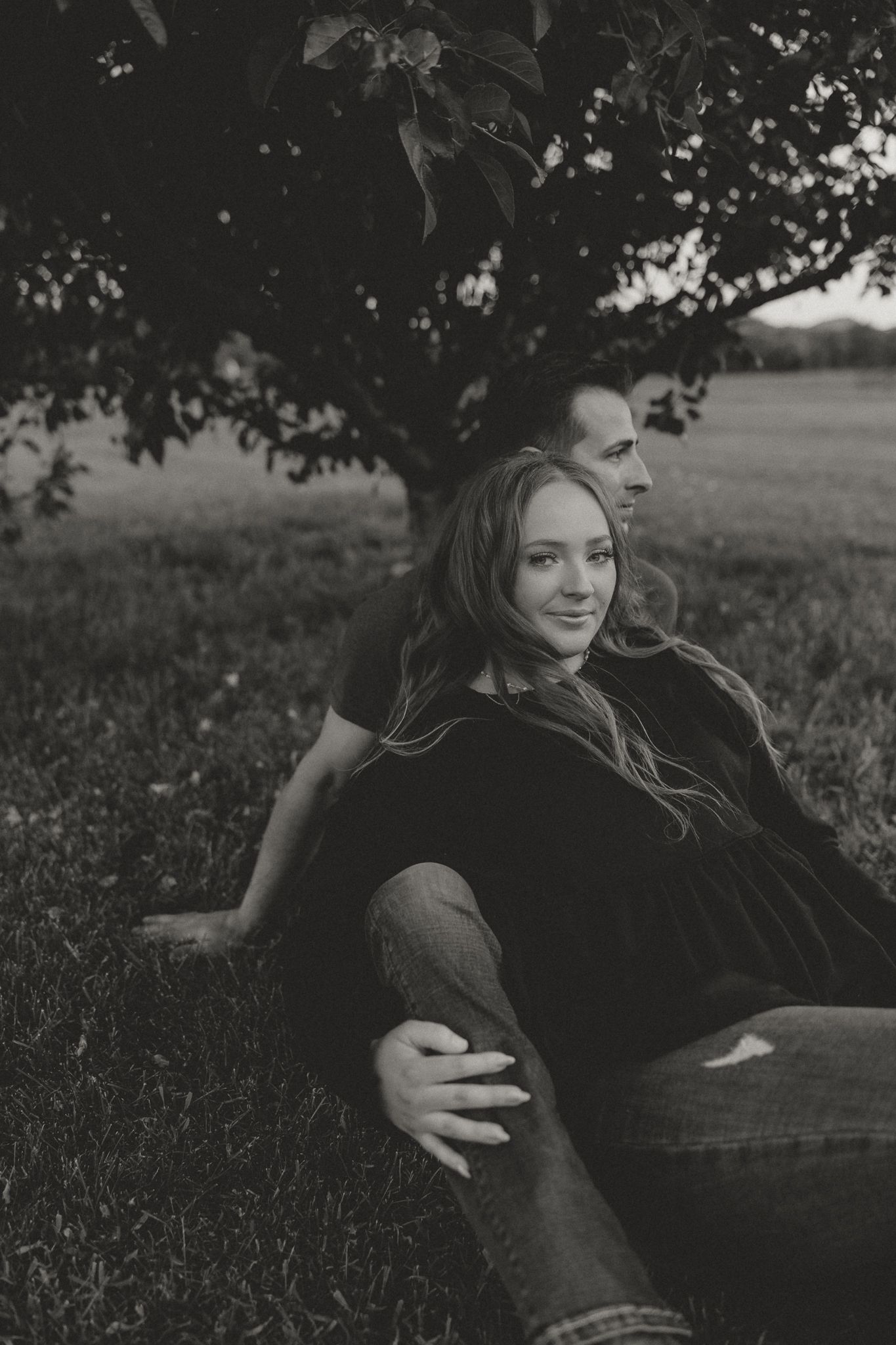A black and white portrait of a couple relaxing under a tree in casual wear.
