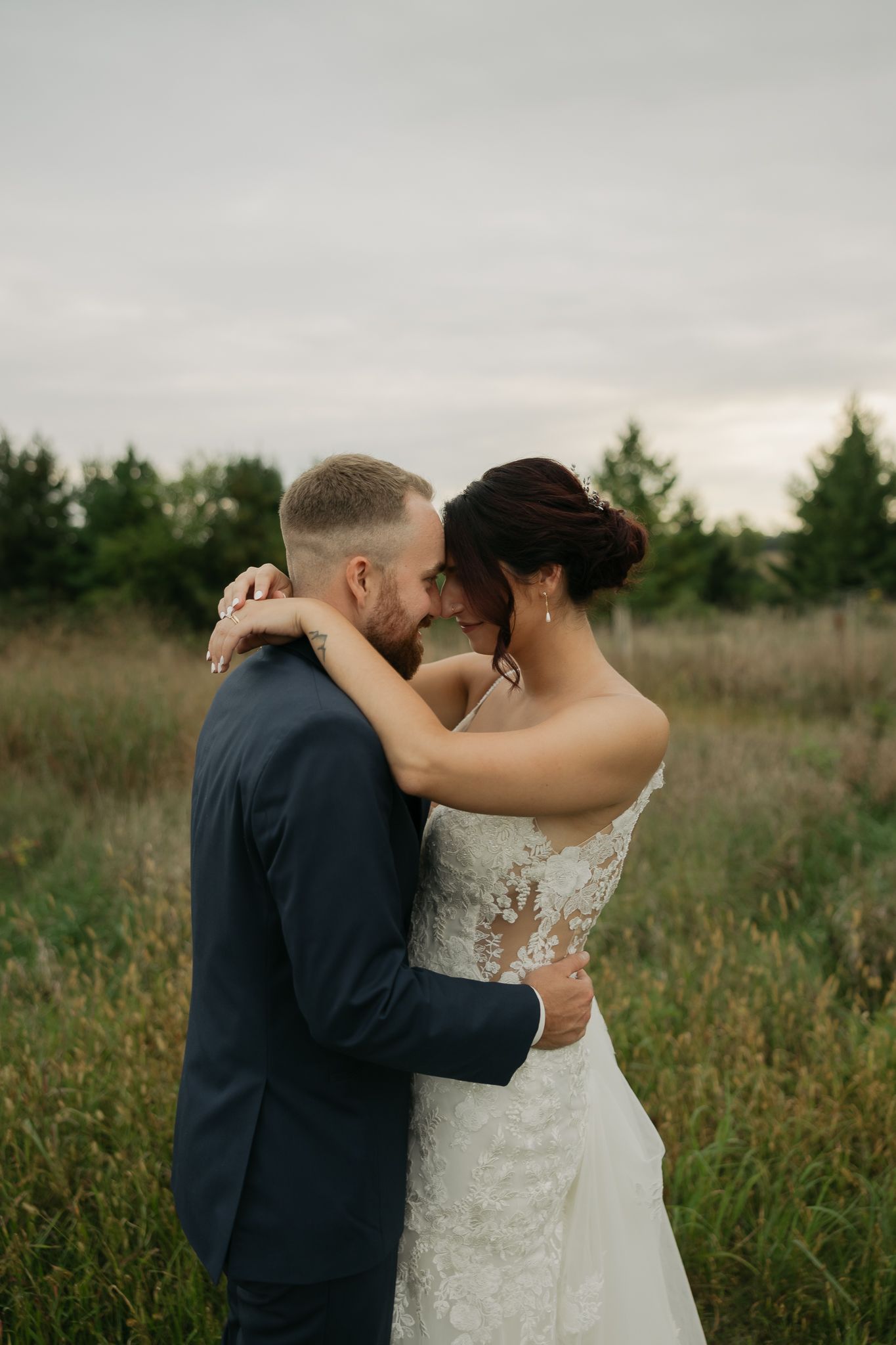 Newlyweds embrace in a field with pine trees and overcast sky in the background.