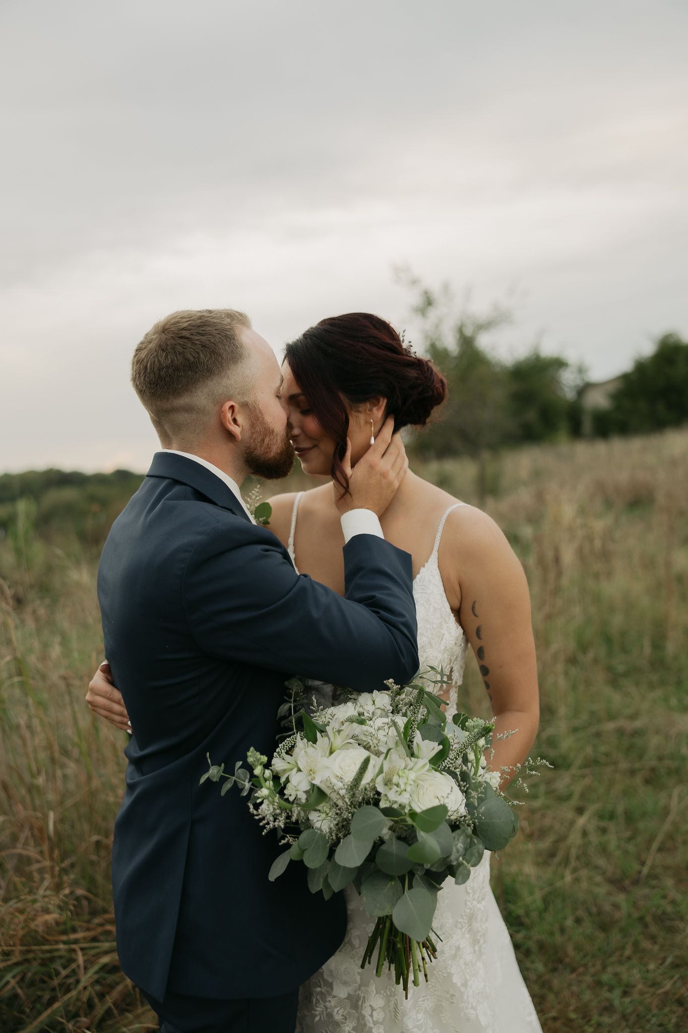 A romantic wedding couple embraces in a field with a white floral bouquet against an overcast sky.