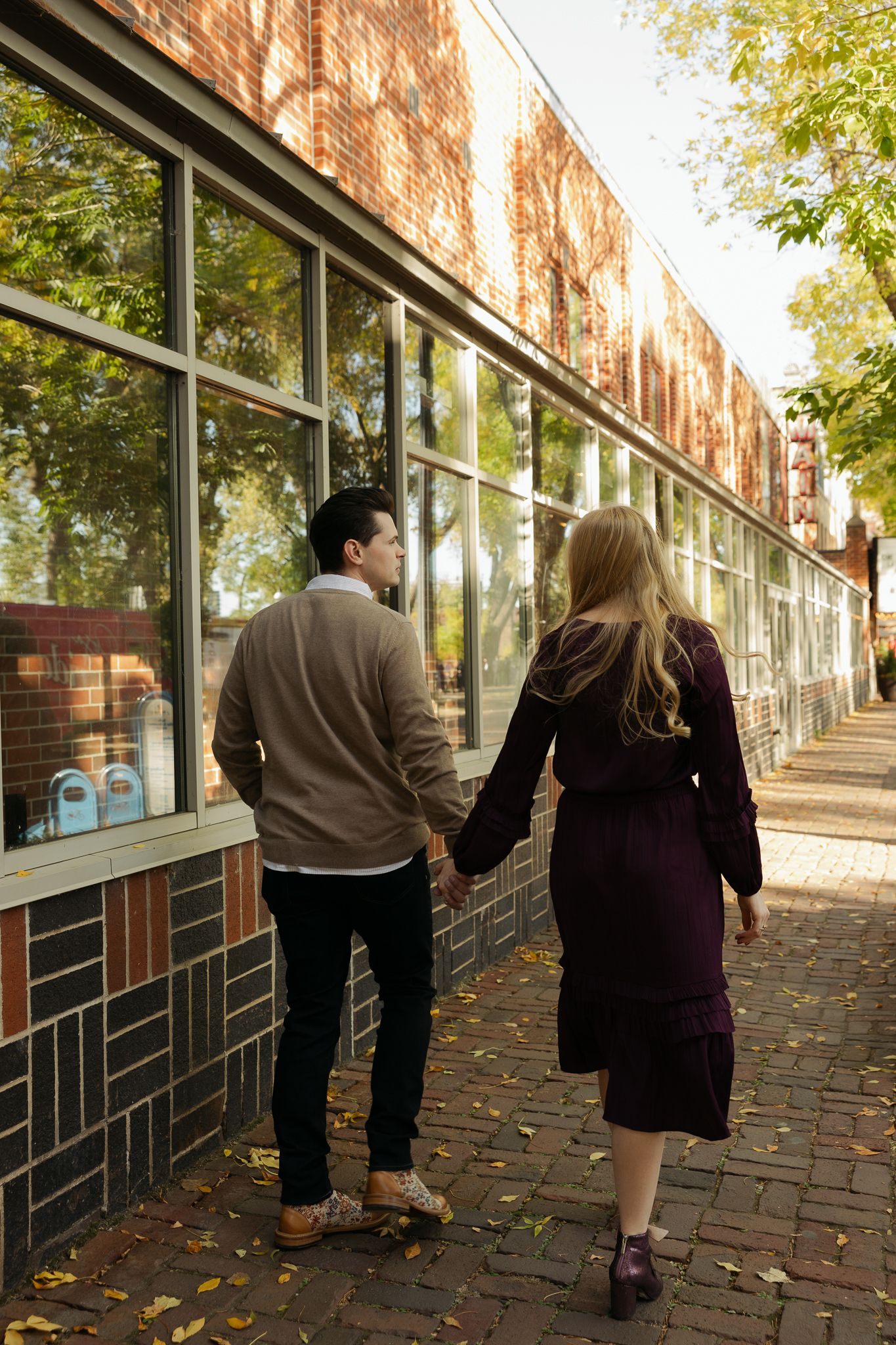 A couple walking together along a brick building with large windows on an autumn day.