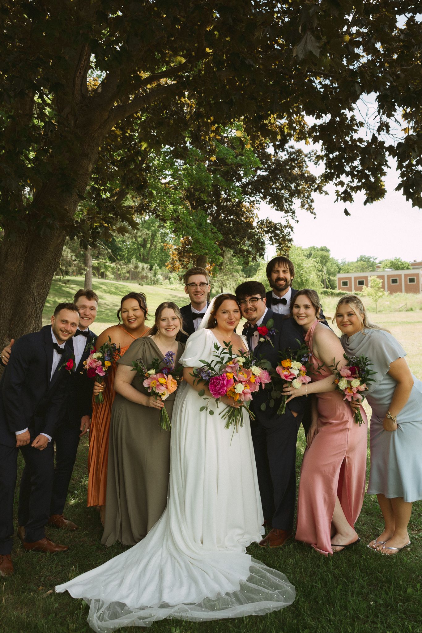 Wedding party poses under a large tree, bride in white gown with colorful bouquets, bridesmaids in earth tones and pink.