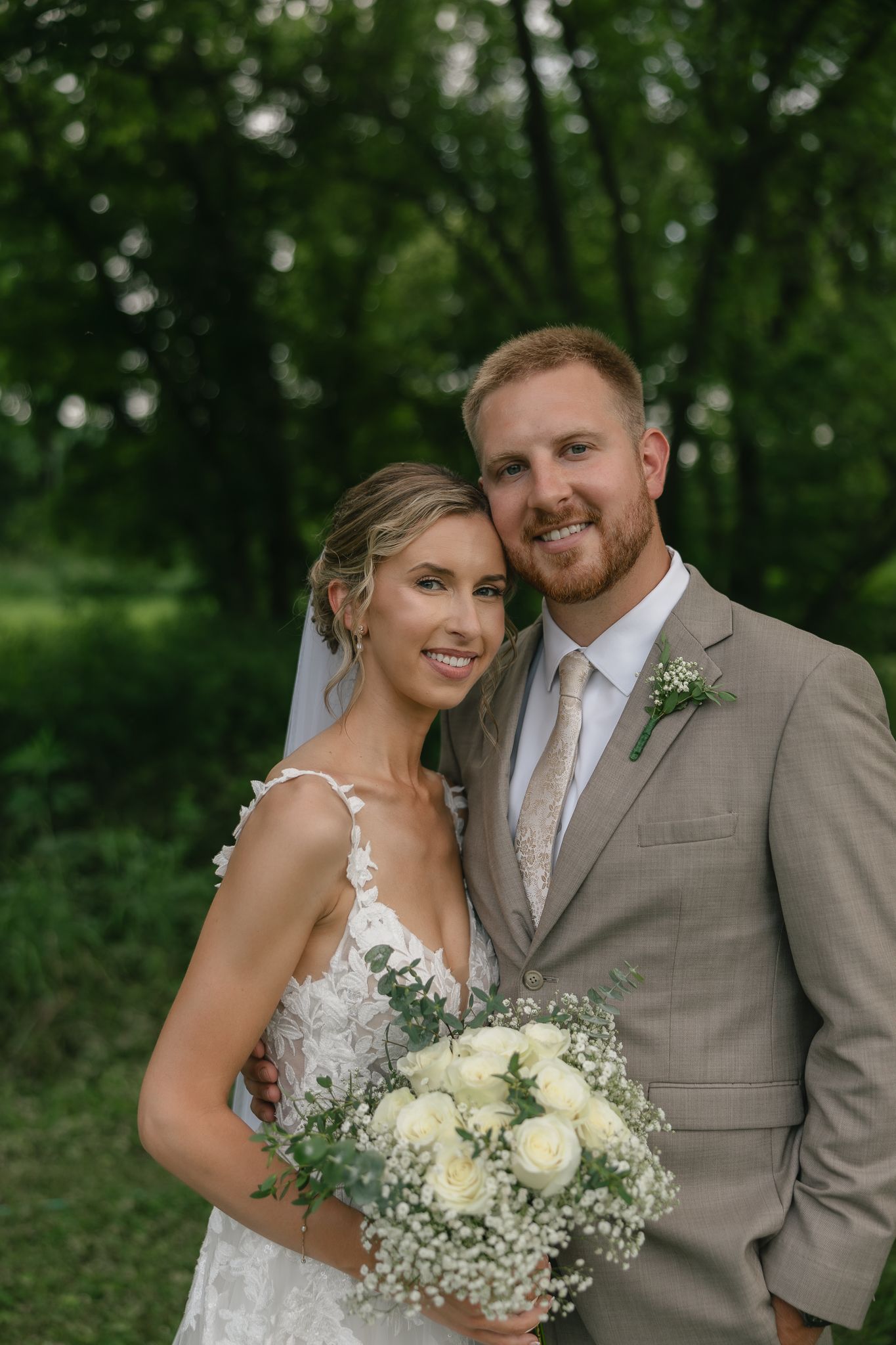 A wedding couple poses outdoors with a beautiful white floral bouquet against a green background.