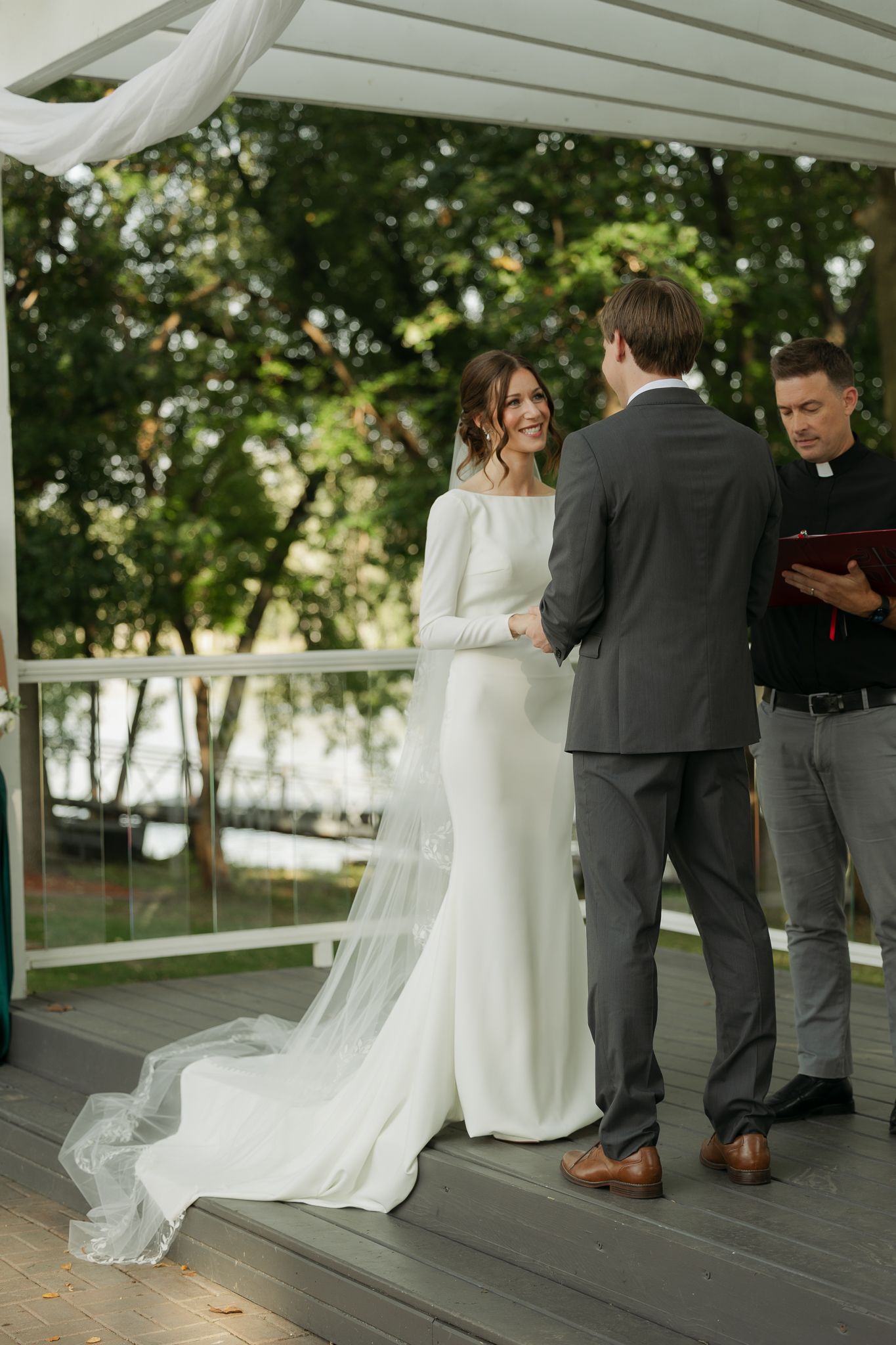 A wedding ceremony takes place under a white pergola with greenery in the background.