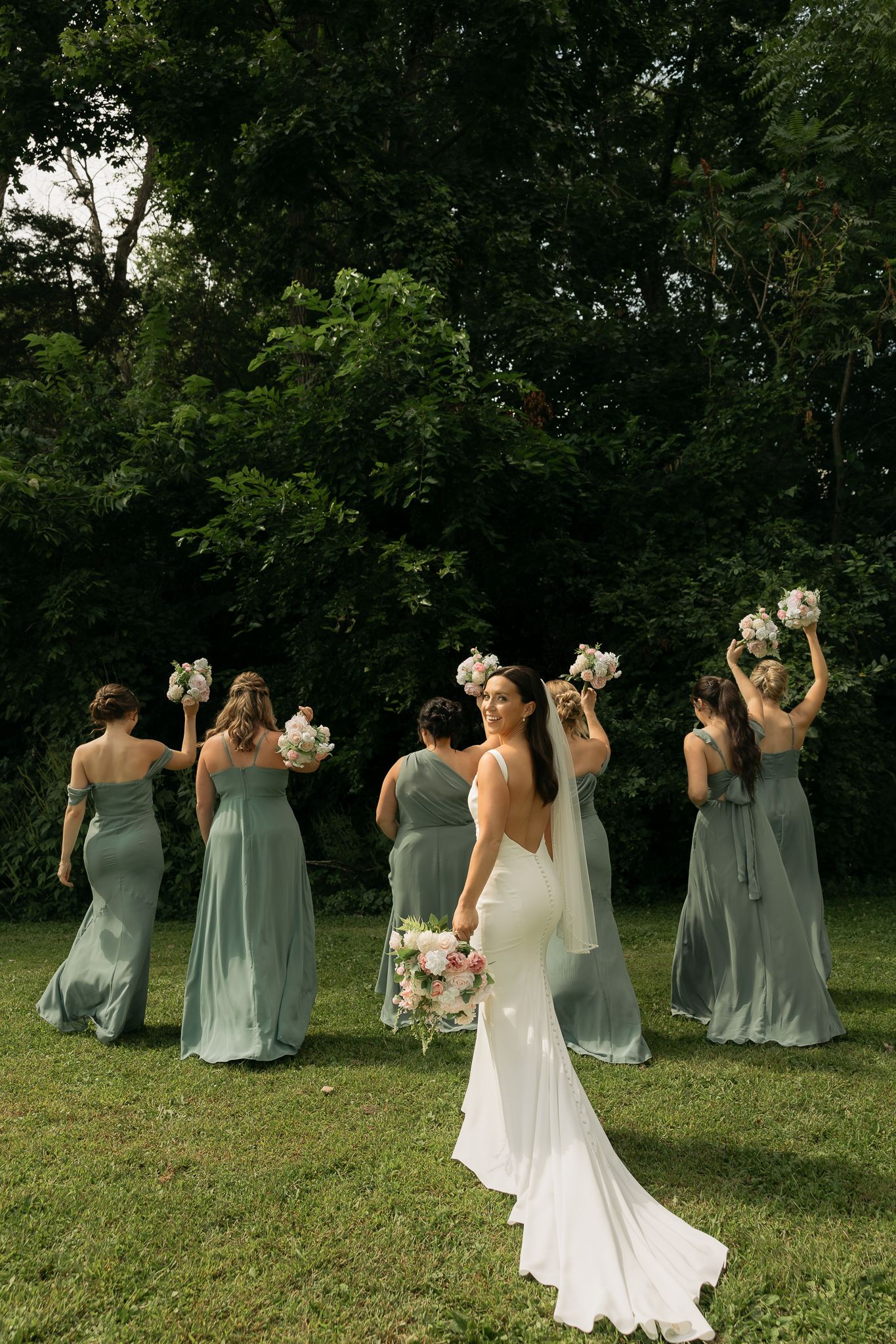 Bride in flowing white gown poses with bridesmaids in sage green dresses holding bouquets in garden setting.