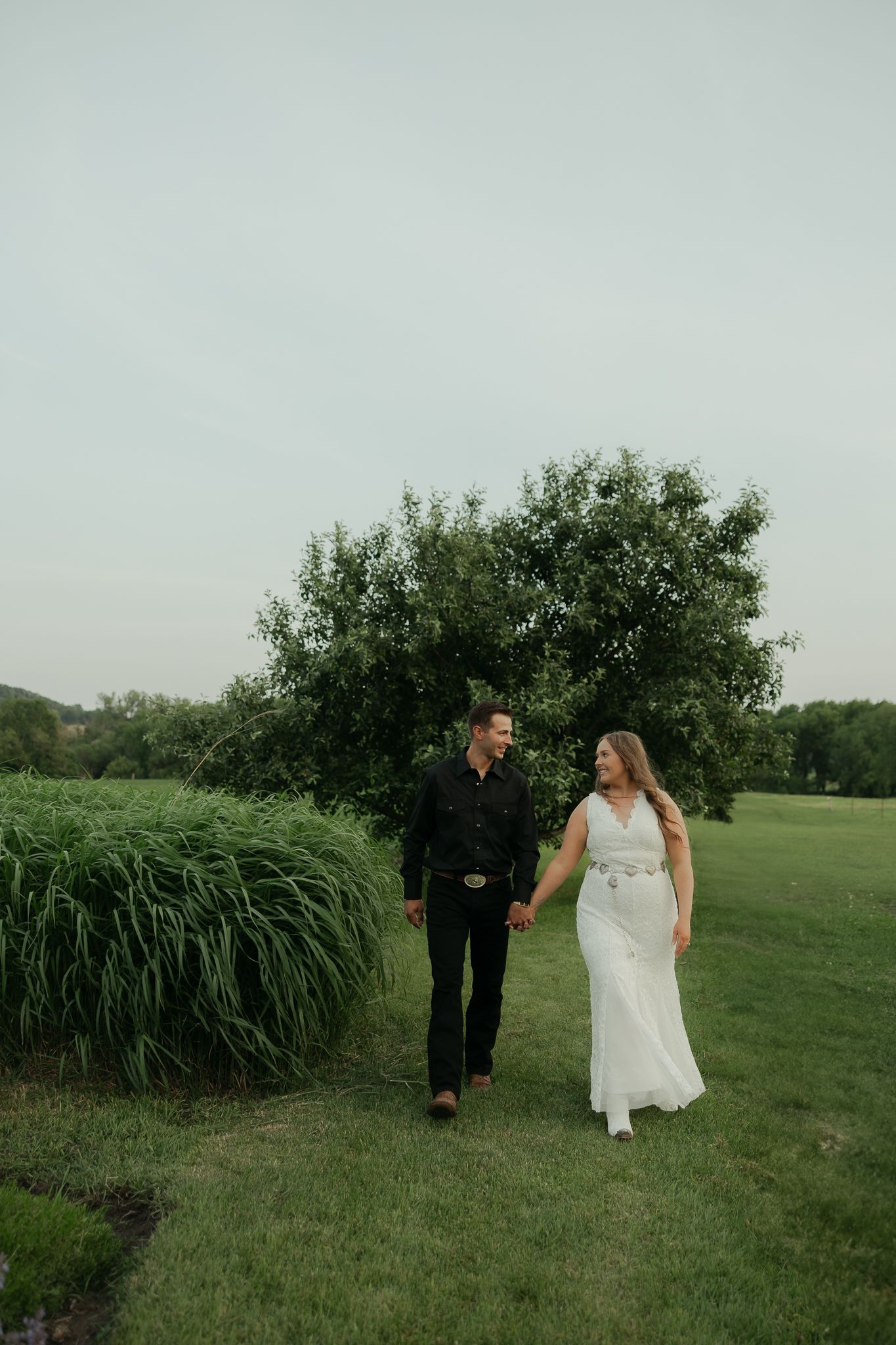 A couple walks together on a grassy path with trees and bushes in the background.