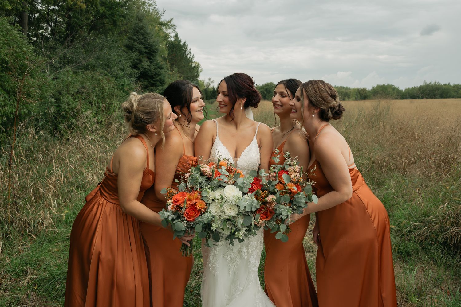 Bridal party with rust-colored dresses gather together holding floral bouquets outdoors.