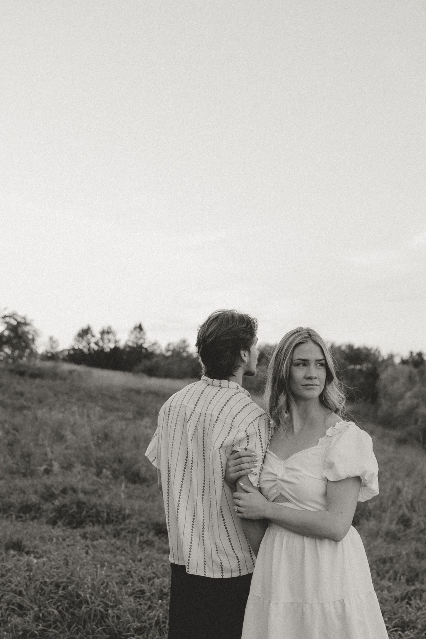 A couple in light colored clothing stands together in a rustic outdoor setting.