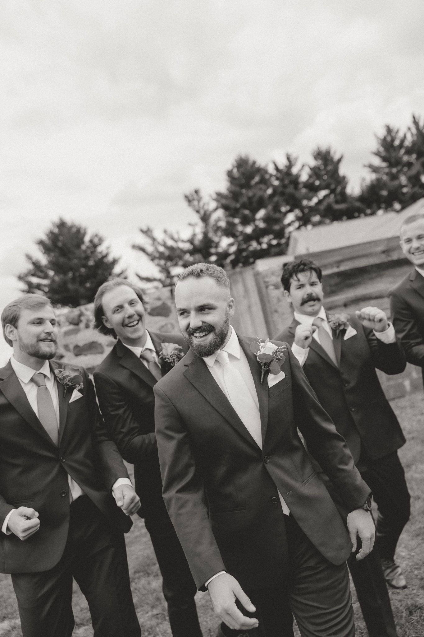 Groomsmen in dark suits laugh together during outdoor wedding photos.