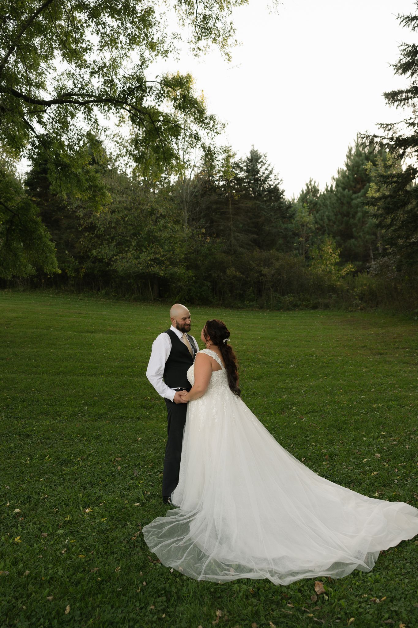 Couple in wedding attire posing on a grassy hillside with flowing white dress trailing behind.