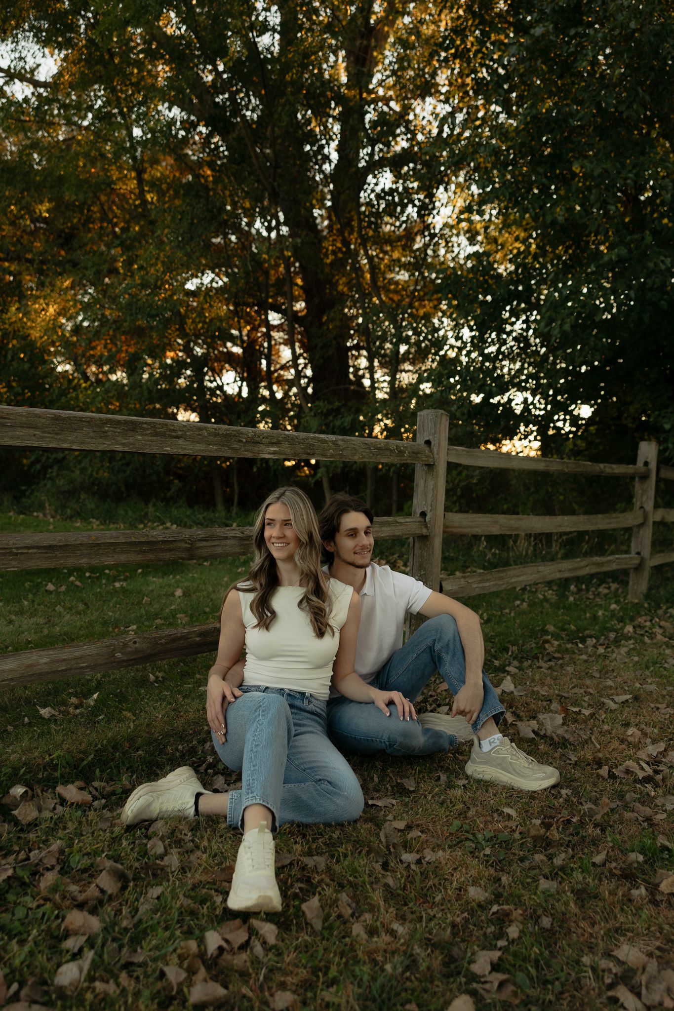 Autumn engagement session with couple relaxing on fallen leaves by wooden fence.