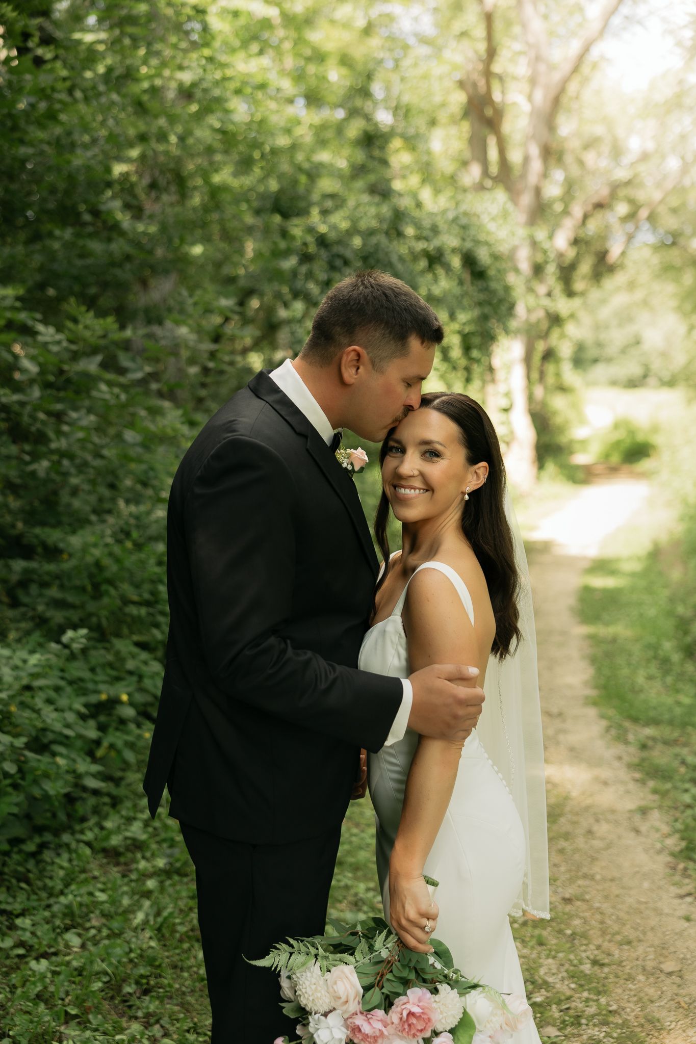 Joyful wedding couple sharing an intimate moment on a sunlit forest path.