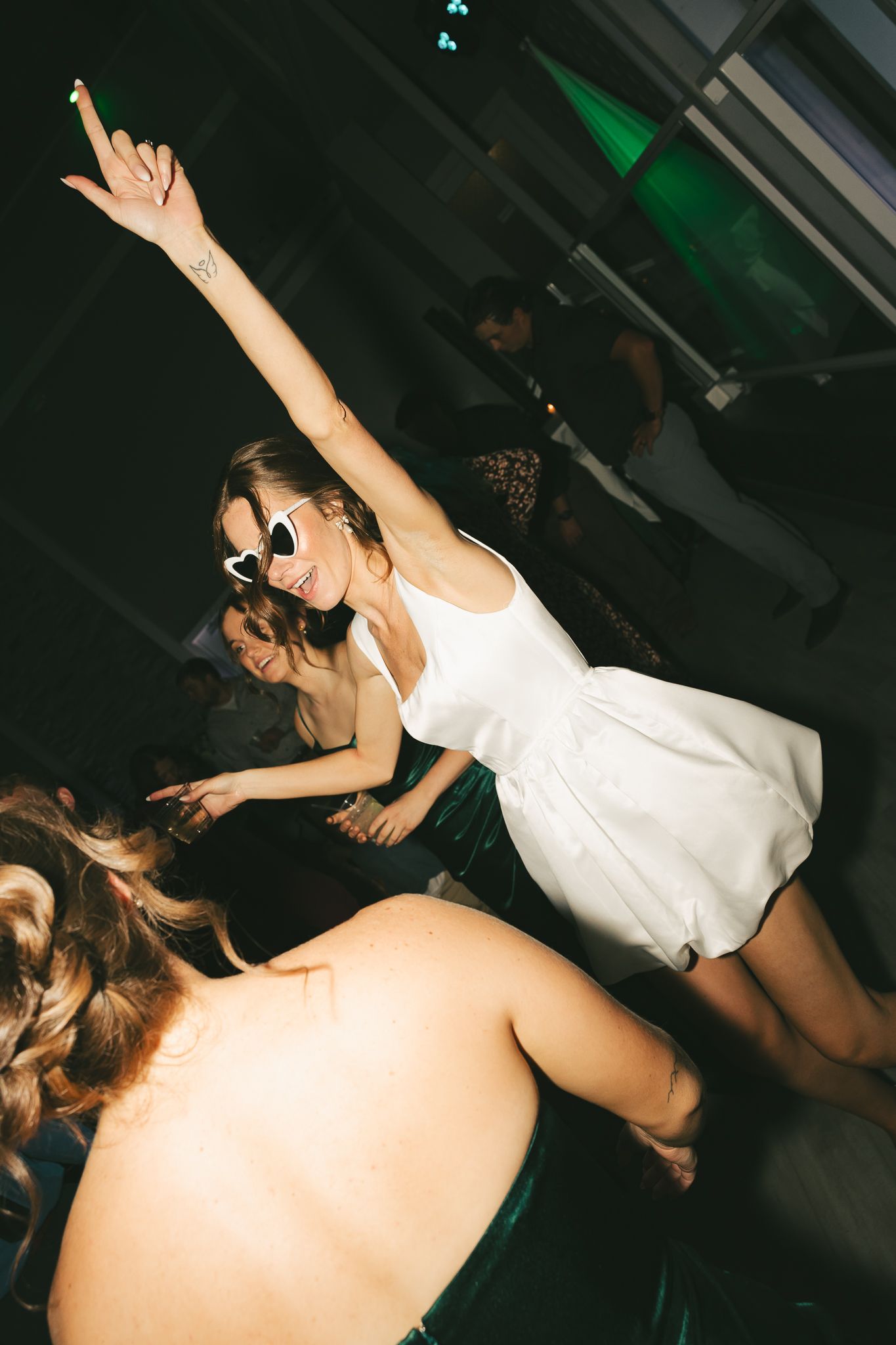 Partygoer in white dress and sunglasses dancing with raised arms on dark dance floor.