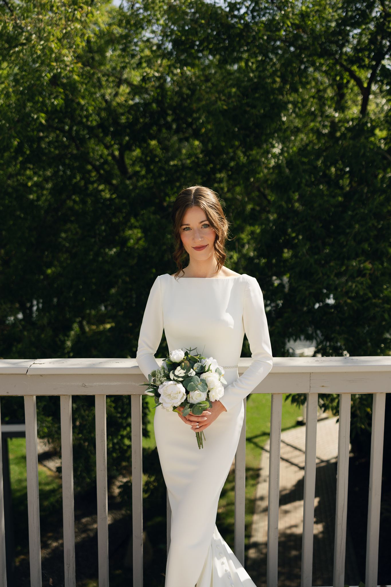 Elegant bride in minimalist white gown poses with bouquet against green outdoor backdrop.