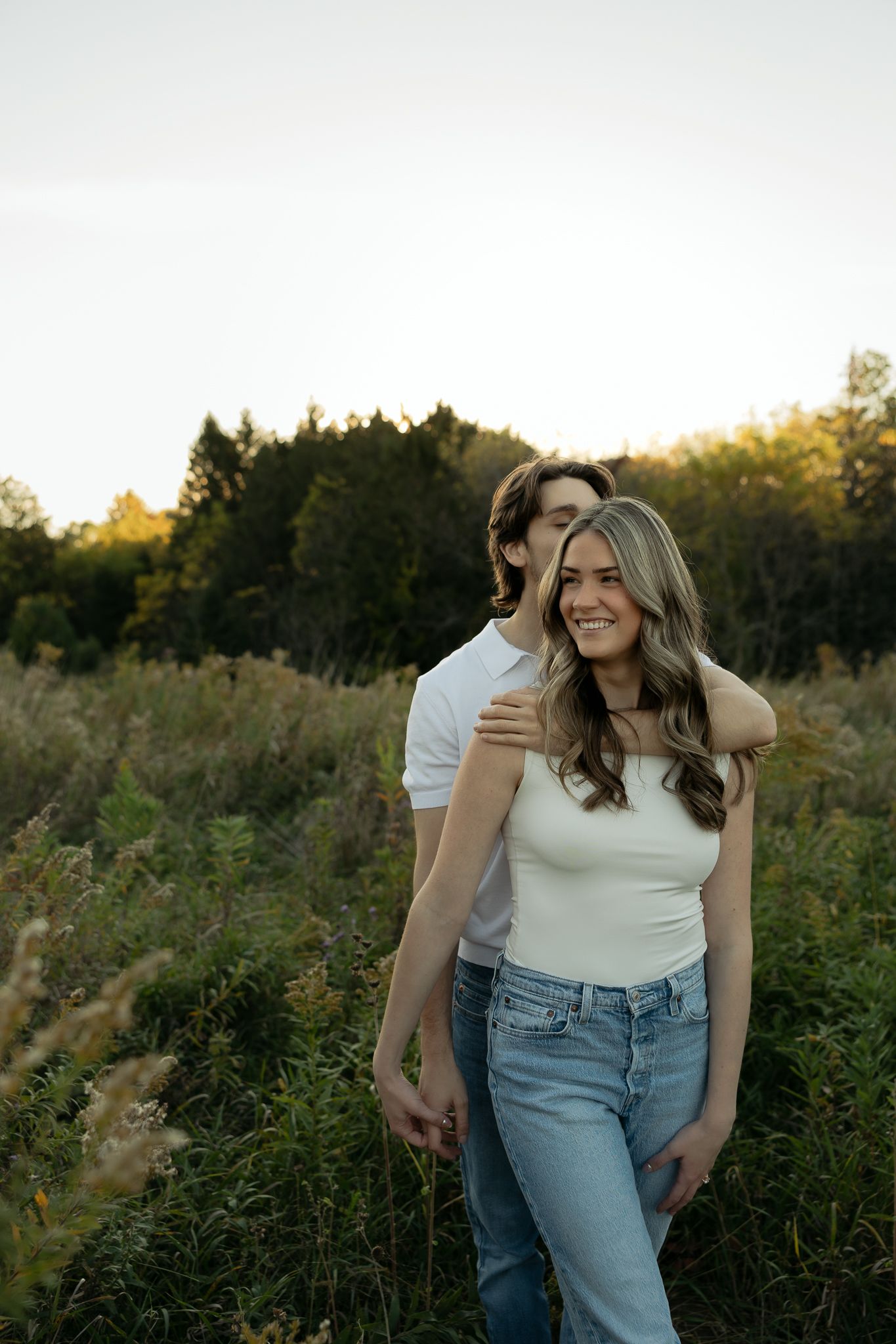 Happy couple in matching white tops and jeans posing together in a meadow at sunset.