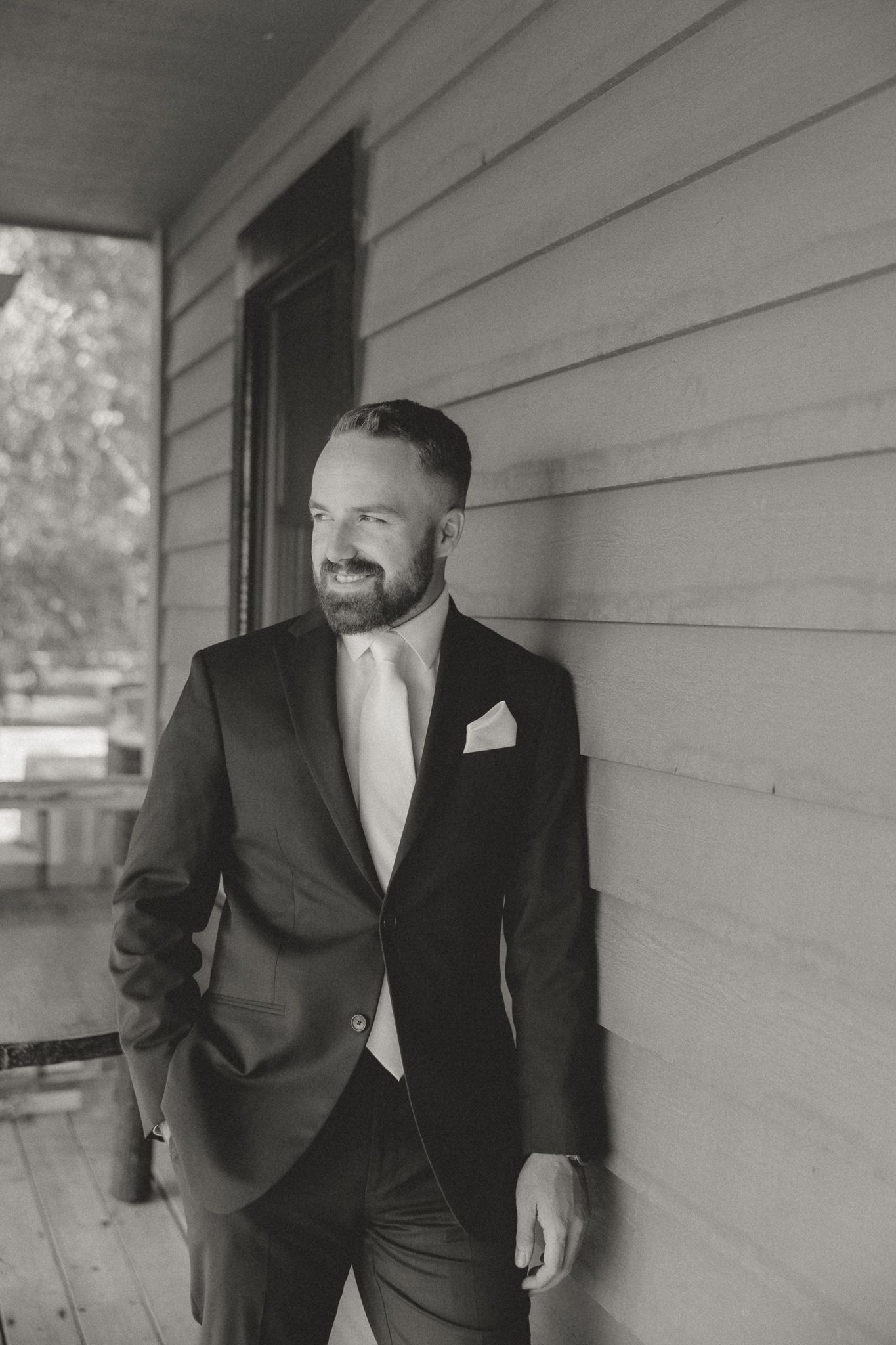Groom in dark suit poses casually against white siding of a building.